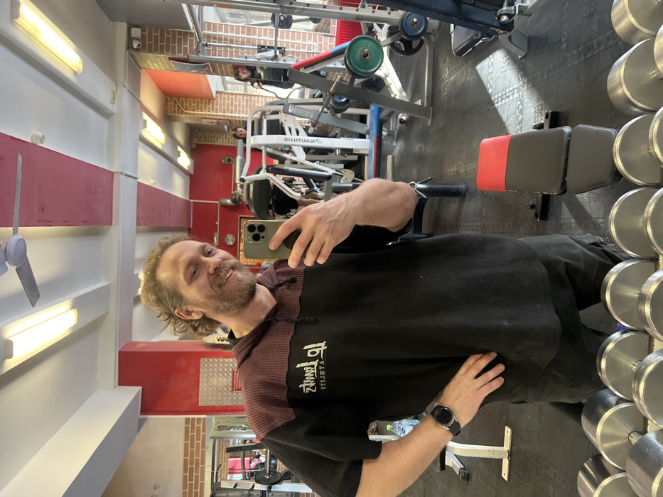 An indoor gym in Saint Petersburg, Russia, is depicted. The primary subject is a male individual with light-brown, curly hair and a beard, smiling directly at the camera while taking a selfie with a grey smartphone held in his left hand. He wears a black and maroon hooded short-sleeved shirt with a mirrored "Up Limits ATHLETE" logo and dark shorts, with a dark smartwatch visible on his left wrist. His right hand rests on his hip.
The foreground shows a rack of chrome dumbbells. In the mid-ground, a black and red adjustable workout bench sits on a dark grey patterned rubber mat floor. The background, largely visible through a mirror reflection, reveals other gym patrons and equipment. To the left, a person in dark attire operates a treadmill. To the right, two women are visible, one on a cardio machine and another standing nearby. Standard gym equipment, including strength machines, weight racks with green, blue, and red plates, and additional benches, populate the space. The walls feature sections of exposed brick, red paint, and lighter panels. The ceiling is fitted with white panels, linear fluorescent lighting, and a white ceiling fan.