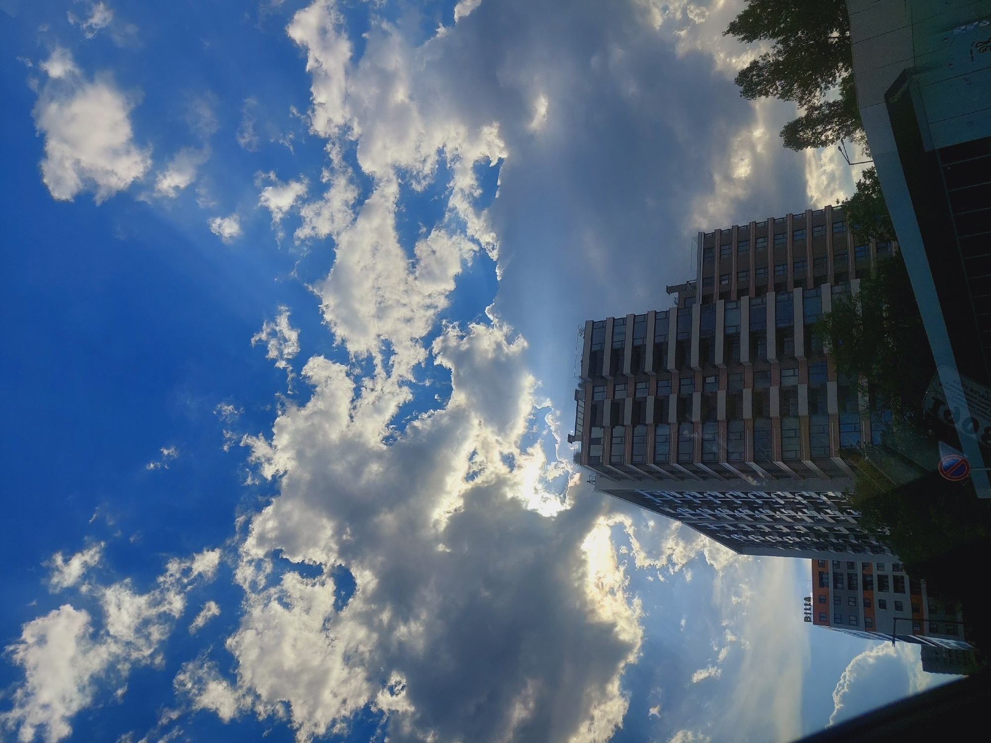 The image shows a modern apartment building in the city of Sofia, Bulgaria. The building is tall and has many windows. It is daytime, and the sky is blue with white clouds. The sun is shining through the clouds, creating a dramatic effect. There are also some trees in the foreground. The building is located in a residential area, and there are other buildings visible in the background. The image appears to have been taken from a low angle, looking up at the building. The overall impression is one of a modern and developing city.