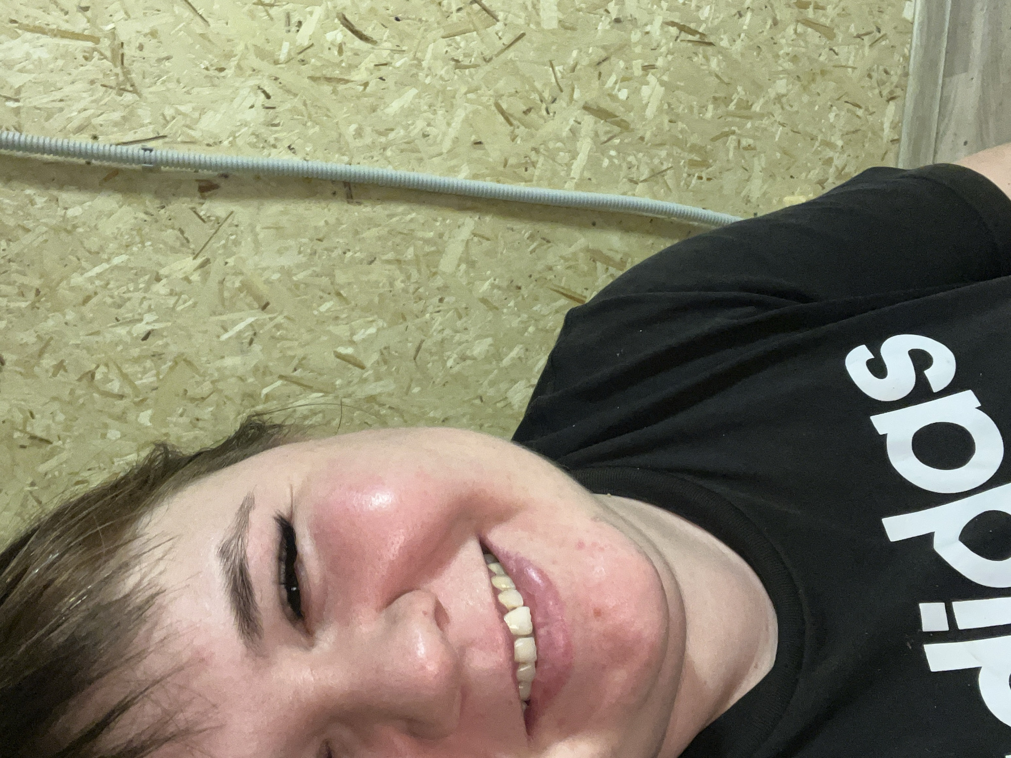 A close-up, angled selfie of a person with fair skin and dark hair, wearing a black Adidas t-shirt. The person is smiling, revealing their teeth. Their face appears flushed, and there are some skin imperfections visible. The background is a wall made of light brown particle board, with a white, corrugated conduit running horizontally across the upper portion of the frame. The location cannot be determined as Chaikovsky, Russia from the given image.