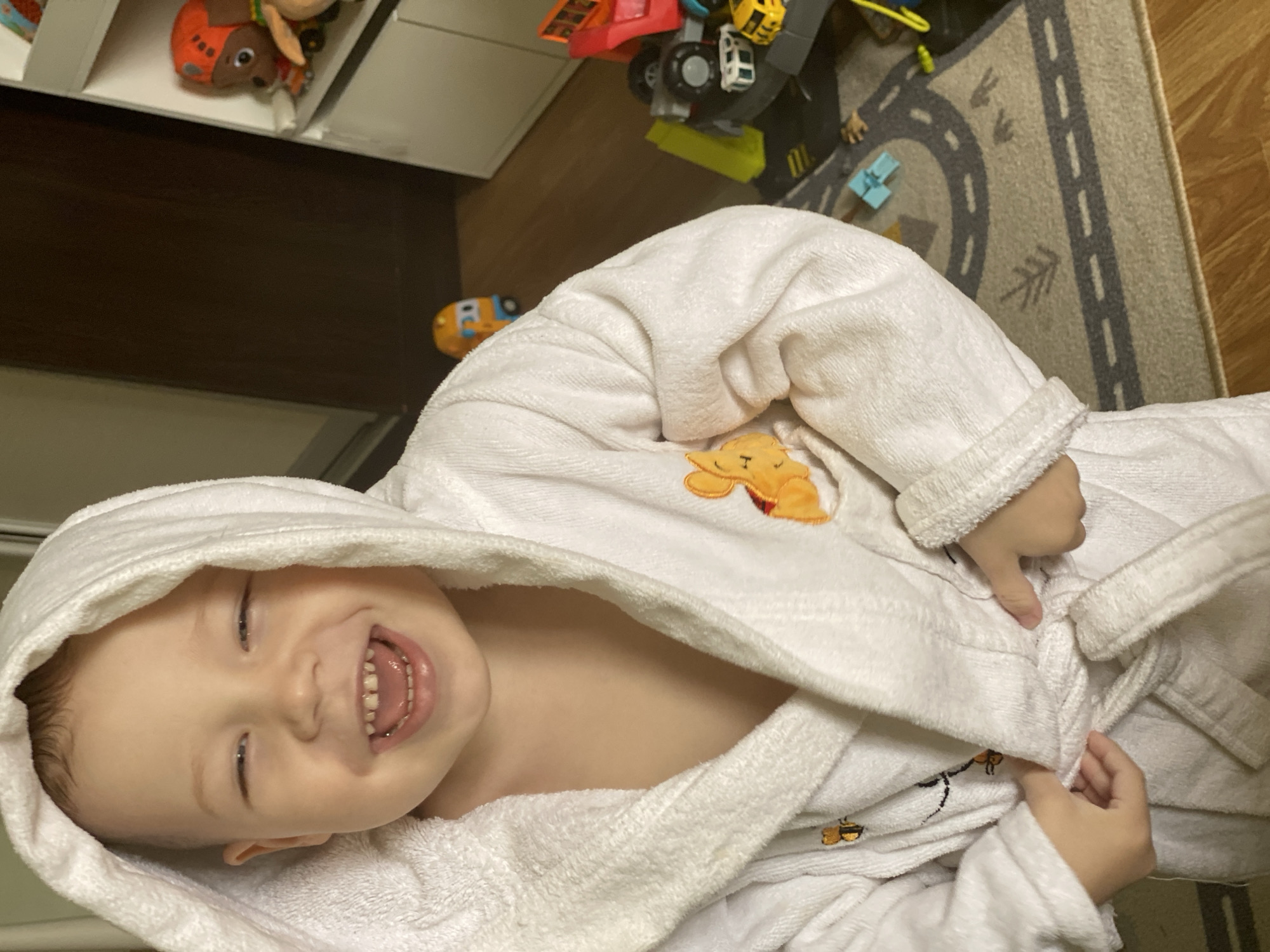 A smiling child wearing a white hooded bathrobe is lying down, likely indoors, in front of some toys and a rug. The surroundings suggest a domestic environment, potentially a child's room. The location of the scene as being in the city of Sochi, Russia, cannot be conclusively confirmed from the visual data.