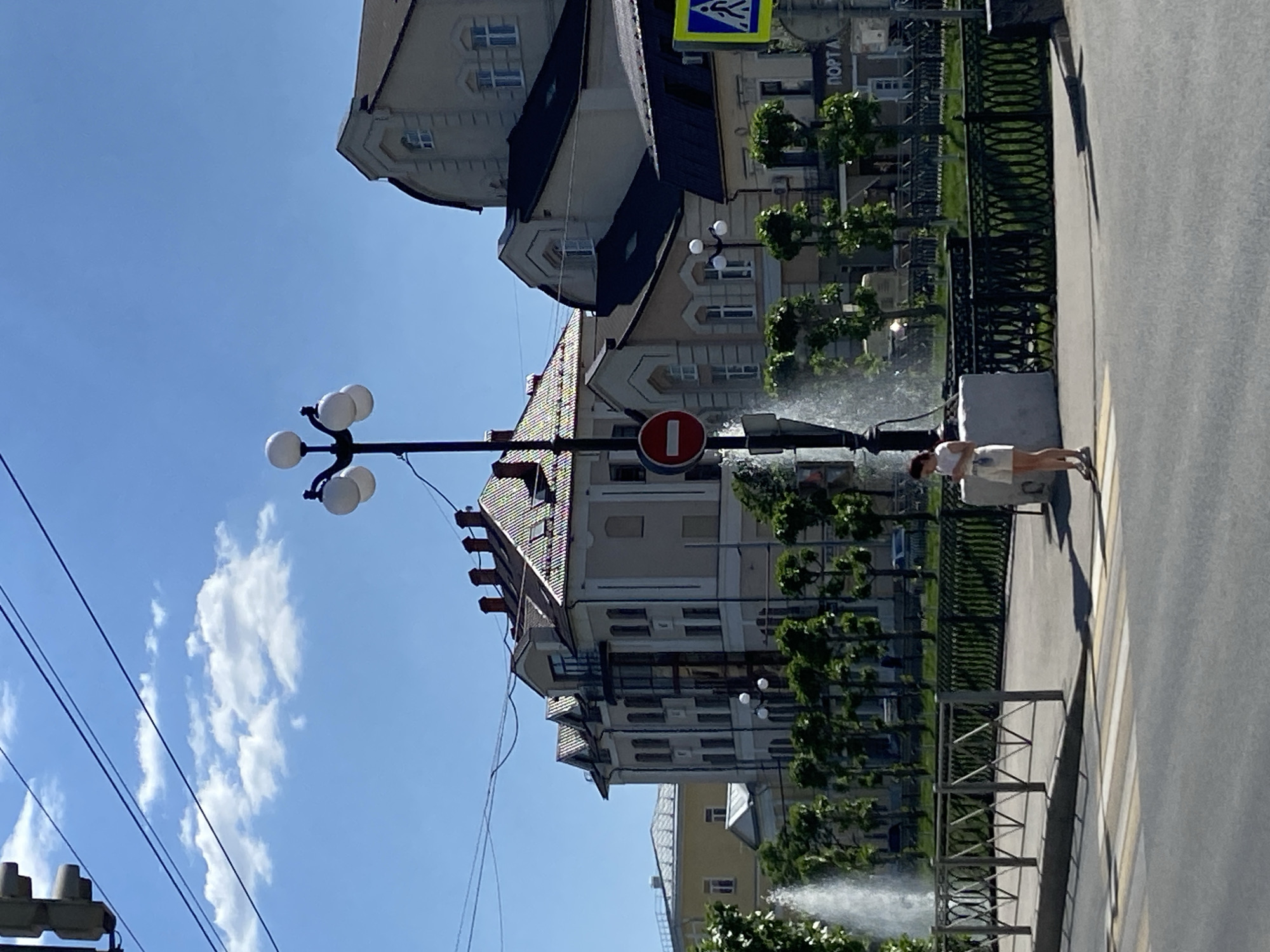 The image shows a street scene in Kazan, Russia, under a clear, blue sky with a few scattered clouds. The buildings are multi-story with varied architectural designs. A street lamp with multiple globe-shaped lights is visible in the left of the frame. A "no entry" sign is prominently displayed on a pole. Water is spraying in the background suggesting a fountain. A person wearing a light colored top and shorts is standing on the sidewalk near a fence. A crosswalk is painted on the road. Power lines stretch across the sky.