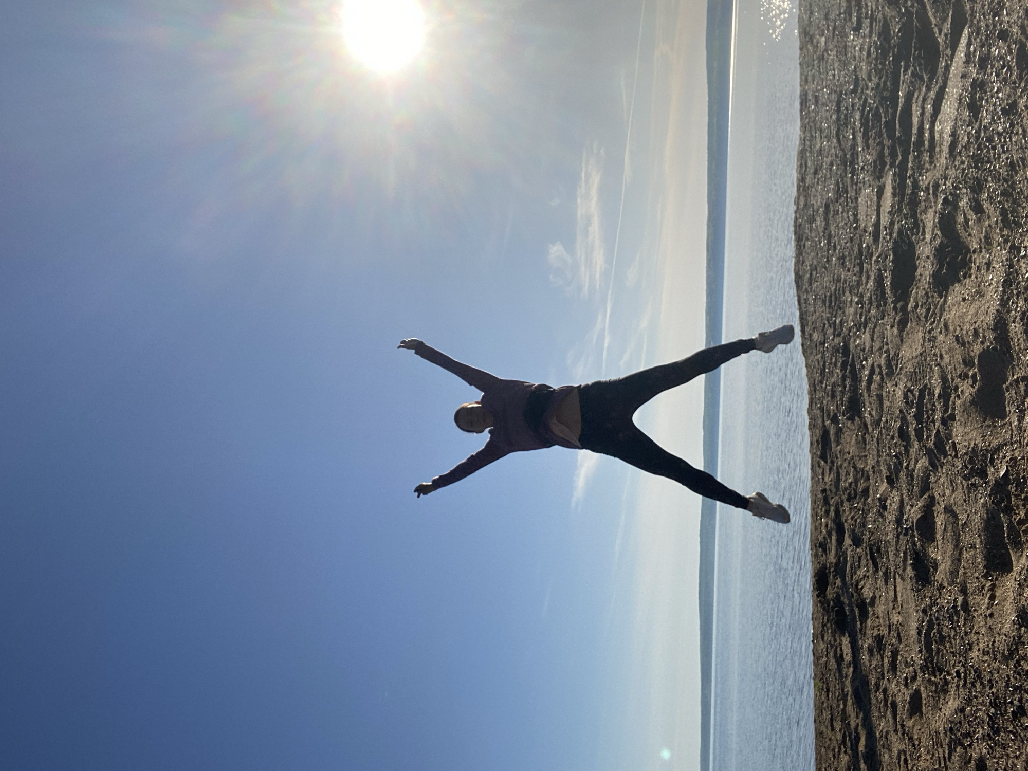 A woman is pictured against a bright blue sky with the sun shining above her. She is positioned with her arms extended outward and legs spread apart, as if mimicking a star or possibly mid-jump. Her attire consists of a jacket, dark pants, and light-colored shoes. The background is divided between the sky and what appears to be a shoreline with water visible in the distance. The shoreline appears to be sandy or rocky. This image was taken in the city of Chaikovsky, Russia.