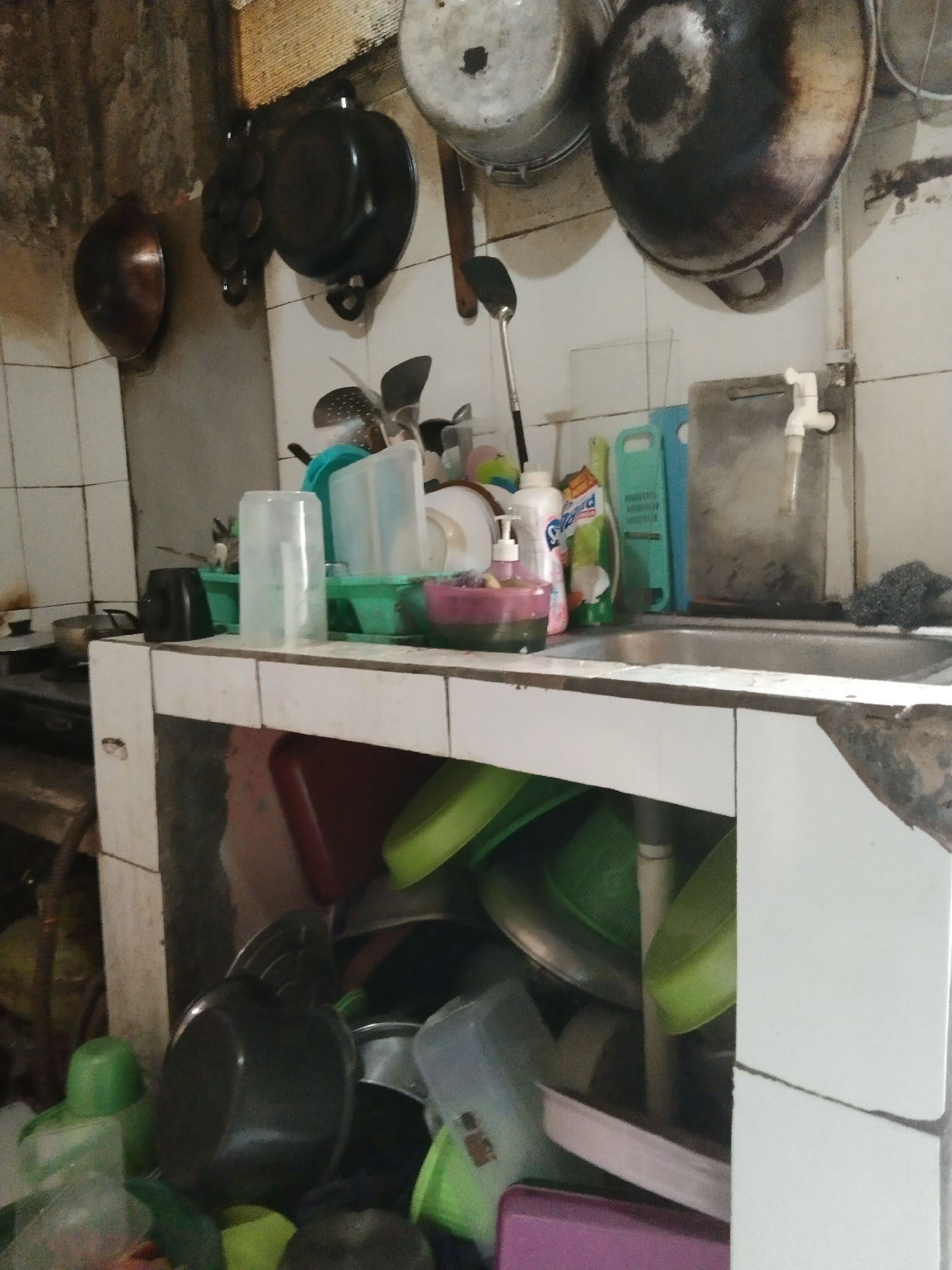 This image depicts a cluttered kitchen in Parung, Indonesia, characterized by disarray and a lack of organization. The scene appears to be inside a residential dwelling.
Dominating the upper portion of the image are several large, dark cooking pots hanging from hooks on a wall. Below these, on a tiled countertop, a variety of kitchen items are piled haphazardly. These include plastic containers of various colors (green, clear, and pink), bottles of cleaning supplies, and what appear to be bowls and other utensils. A stainless steel sink is visible on the right side of the countertop, with a white faucet. A metal scrubber rests next to the sink.
The lower part of the image reveals a storage area beneath the countertop, also filled with numerous items, primarily plastic bowls and containers in green and other colors, along with some darker pots. The overall impression is one of a busy, heavily used kitchen that is in need of cleaning and tidying. The walls are tiled, but some areas show signs of age or disrepair, with staining and grime. The lighting is dim, contributing to the overall sense of a utilitarian and somewhat neglected space. No individuals are present in the frame.