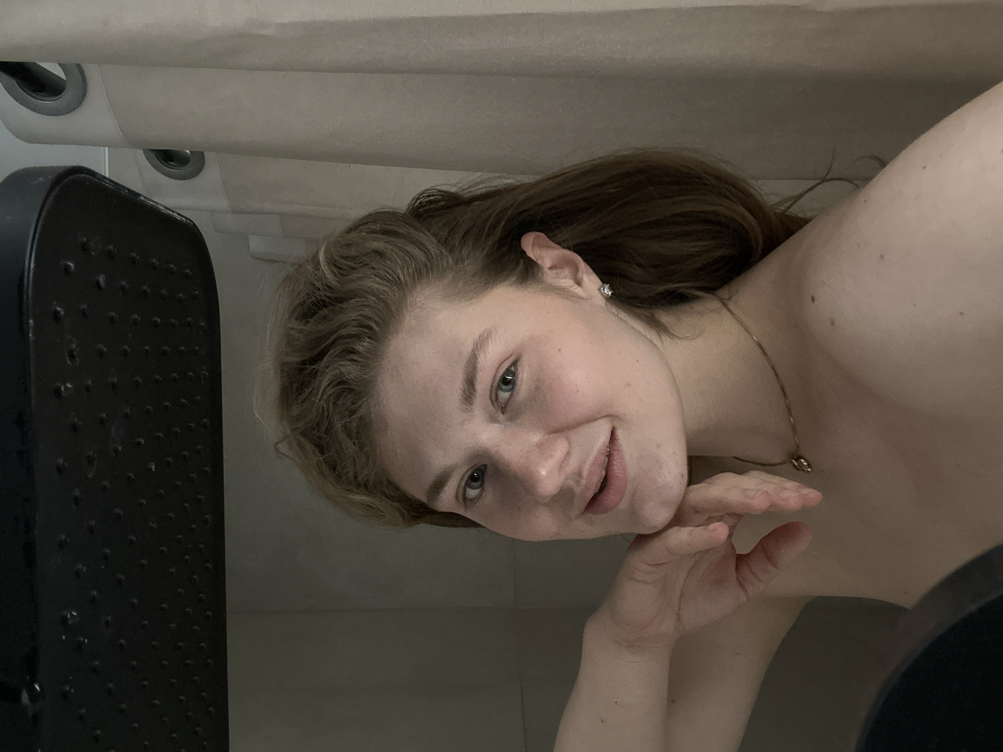 A woman with fair skin and light brown hair is lying down, posing for a selfie or a photo. She is wearing a delicate gold necklace and a small earring. Her hand is gently touching her chin. A black object with a dotted pattern is visible to her left. The background consists of what appears to be an interior with light-colored walls. There is no indication of Kazan, Russia, or any other city.