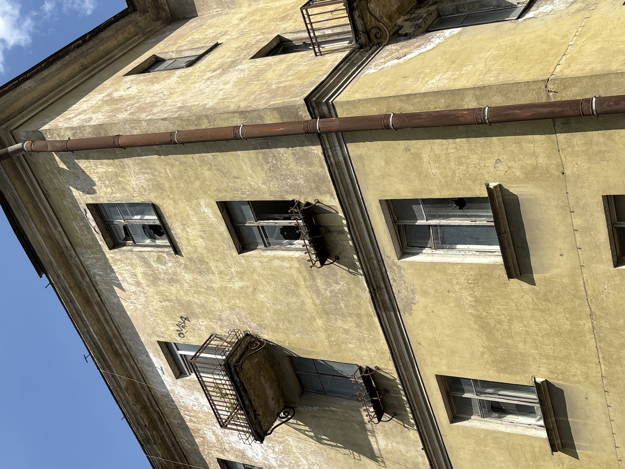 The image shows a close-up, low-angle view of the corner of an old, weathered apartment building in Saint Petersburg, Russia. The building's facade is painted a faded yellow, with visible signs of age such as peeling paint, cracks, and discoloration. A rusty brown downspout runs vertically along the corner of the building.
There are several windows visible, some with broken panes, indicating disrepair. One window has a small, black object inside, possibly a silhouette of a cat. Two balconies are also visible, both with ornate, rusty metal railings. One balcony has a small plant growing in a pot, while the other appears empty. On the upper part of the building, faint graffiti in black spray paint can be seen on the wall, appearing to spell out "OMSA". The sky above is a clear, bright blue with a few wispy white clouds. The overall impression is one of urban decay and neglect.