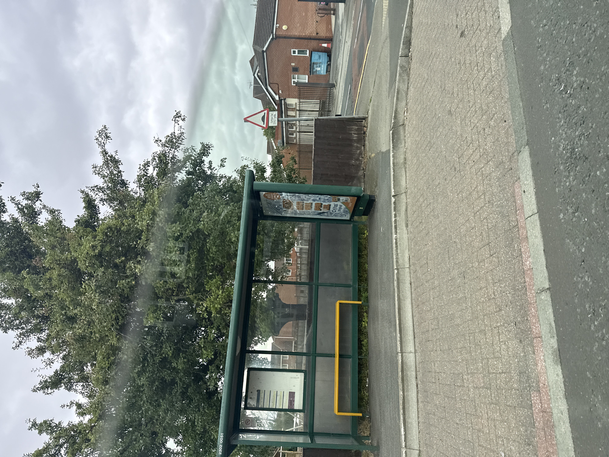 The image shows a bus stop in Darlington, United Kingdom, under a cloudy sky. The bus stop has a green frame with glass panels and a yellow bench. A timetable is visible inside the bus stop. To the right of the bus stop, there is a brick building with a fence and a blue bin. A speed bump sign is also present. The ground is paved with brickwork. The overall scene is typical of a residential area in the UK.