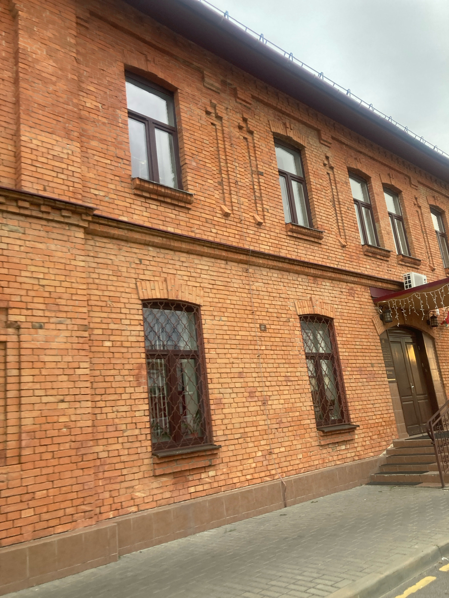 The image shows a brick building with multiple windows. The building is located in the city of Brest, Belarus. There are no people or animals visible in the image. The building has a red brick facade and brown window frames. Some of the windows have security bars. There is a sidewalk in front of the building, made of grey pavers. The sky is overcast.