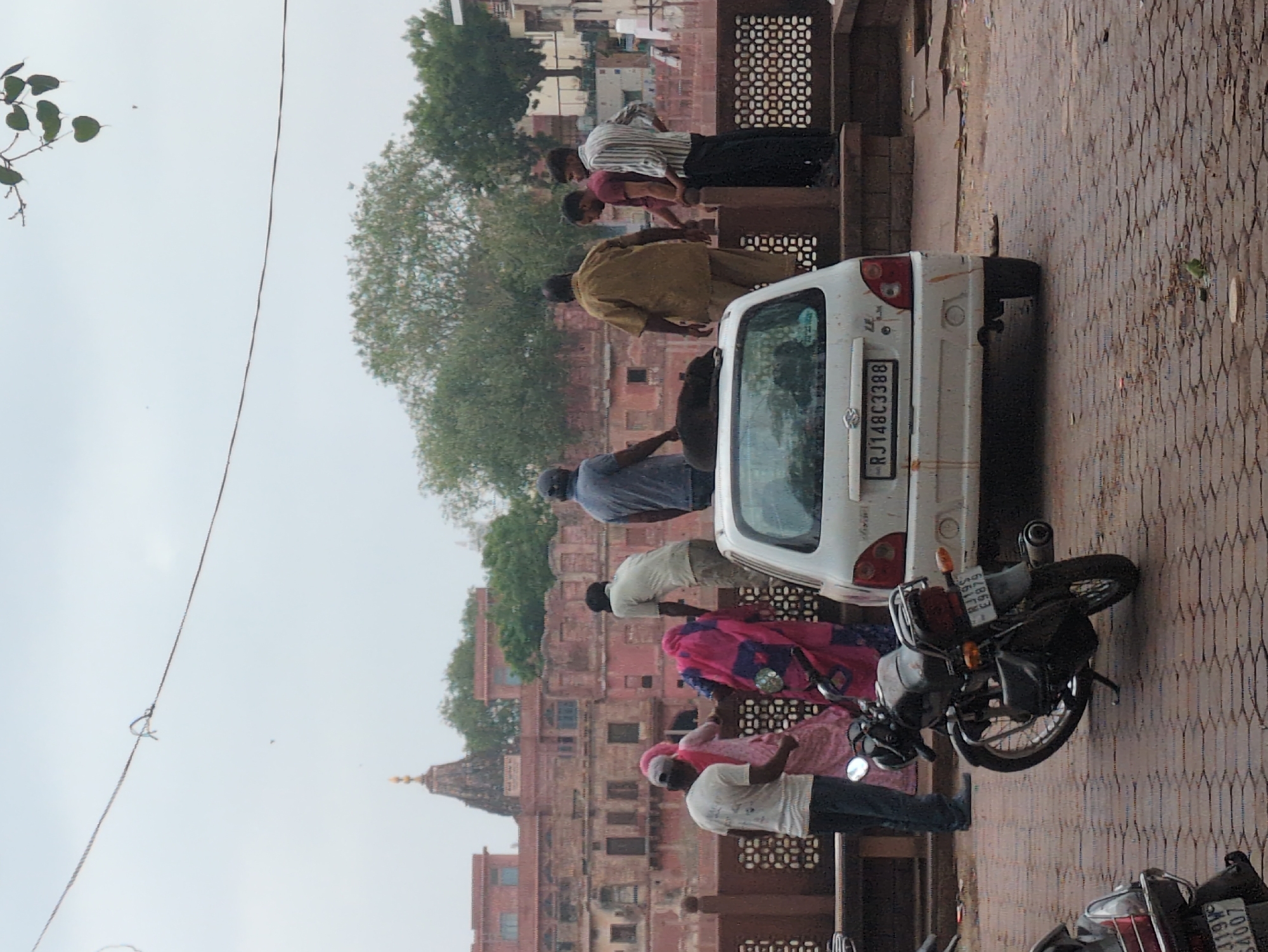 The image depicts a street scene in Jodhpur, India, with a temple structure visible in the background. In the foreground, a white car is parked, and a black dog is lying on its roof. Several people are standing around the car and on a raised platform behind it. A motorcycle is parked to the left of the car. The ground is paved with hexagonal stones. The sky is overcast, suggesting a cloudy day. The architecture in the background, characterized by its red sandstone construction and ornate detailing, is consistent with the style found in Jodhpur.