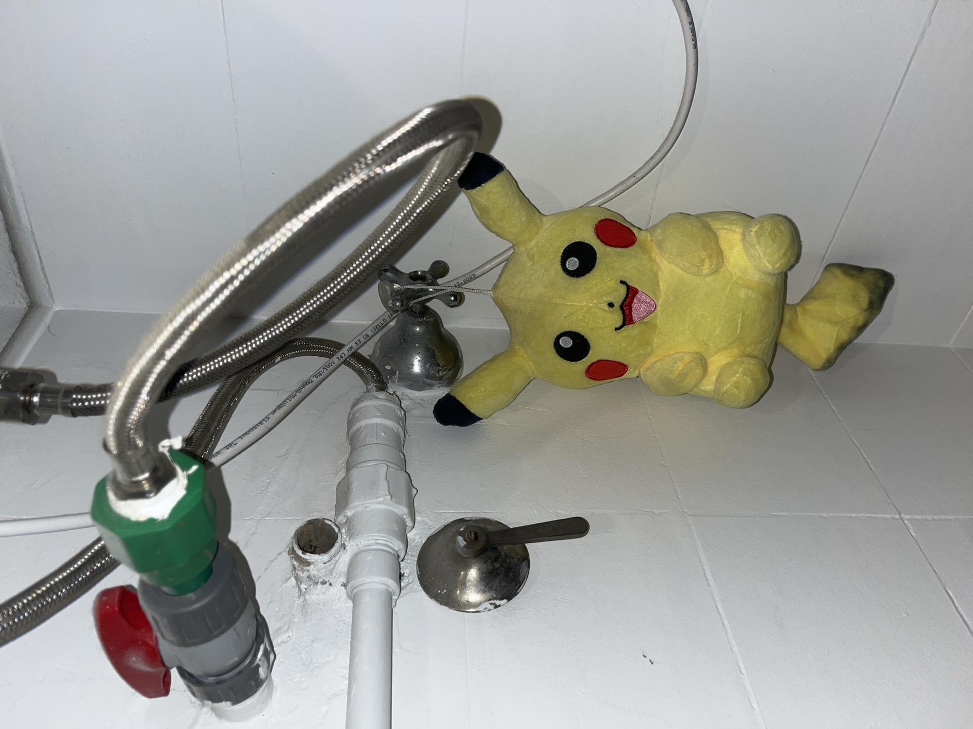 The image shows a close-up view of a plumbing fixture under a sink, with a Pikachu plush toy lying beside it. The plumbing consists of braided stainless steel flexible hoses connected to PVC pipes and shut-off valves. A white electrical cable runs overhead. The background consists of a white tiled floor and wall. The location is not indicated in the image.