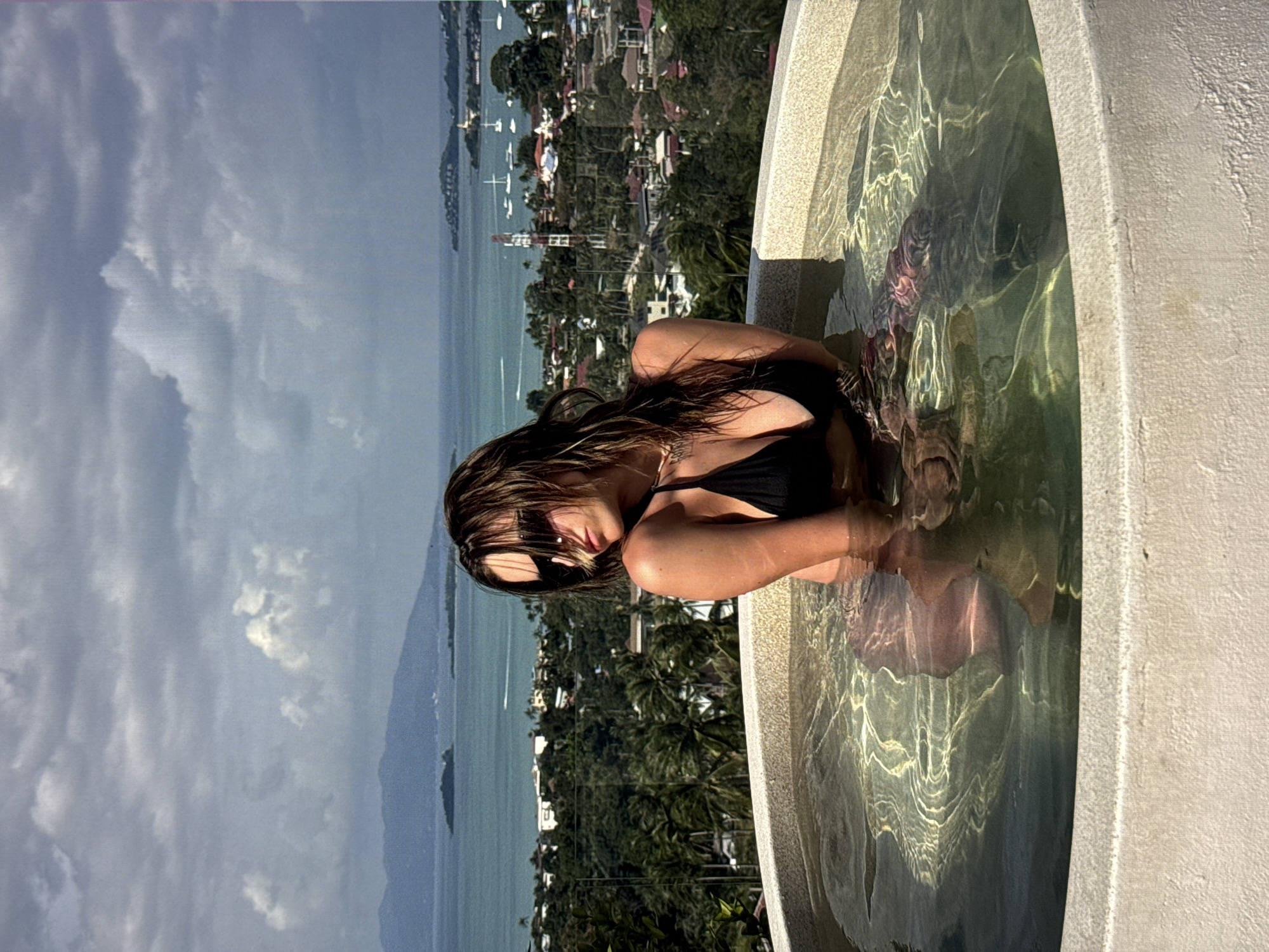 A woman in a black bikini is submerged in a circular jacuzzi with a textured, light-colored rim. She is wearing sunglasses and has wet hair. The jacuzzi is positioned outdoors with a scenic background of a coastal city and islands under a cloudy sky. The city appears to be built on hills with a mix of houses, vegetation, and a small harbor with boats. The image does not contain any elements that suggest the location is Moscow, Russia.