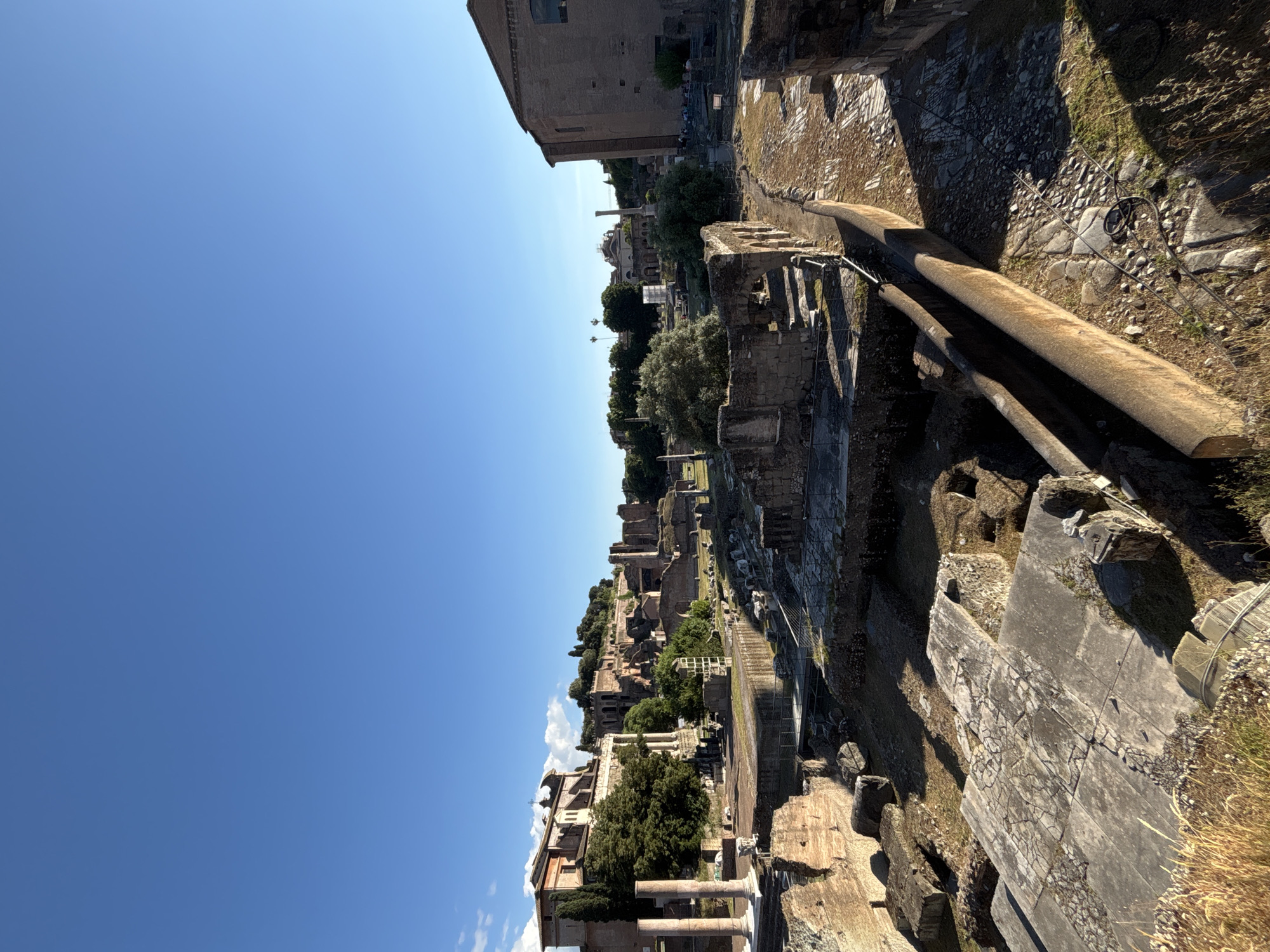 The image shows the ruins of the Roman Forum in Rome, Italy, on a clear, sunny day. The camera angle is slightly elevated, looking down on the ruins. In the foreground, weathered stone structures and architectural fragments are visible. Further into the scene are more ruins, trees, and vegetation. In the distance are other buildings and structures within the city. The sky is a clear blue, with no visible clouds. There are no visible people in the image.