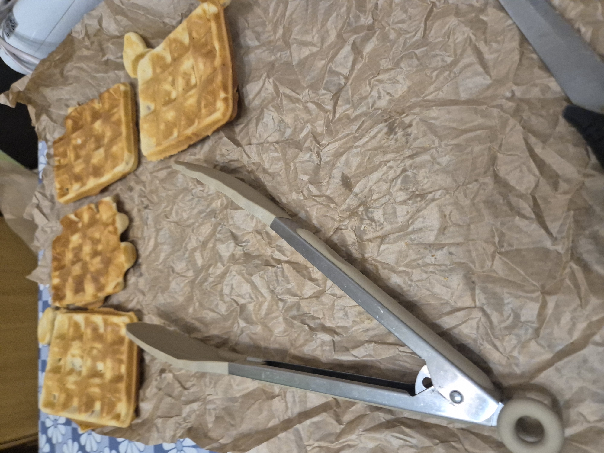 The image shows a close-up view of five waffles on a sheet of crumpled brown paper. The waffles are golden-brown and have a square patterned texture. A pair of metal tongs is positioned diagonally across the center of the paper. The handles of the tongs are grey with beige rubberized tips. In the upper left corner of the image, part of a white appliance is visible. In the upper right corner, a black handled knife is present. A blue and white floral surface is visible on the left and bottom edge of the image. The location is not evident in the image.