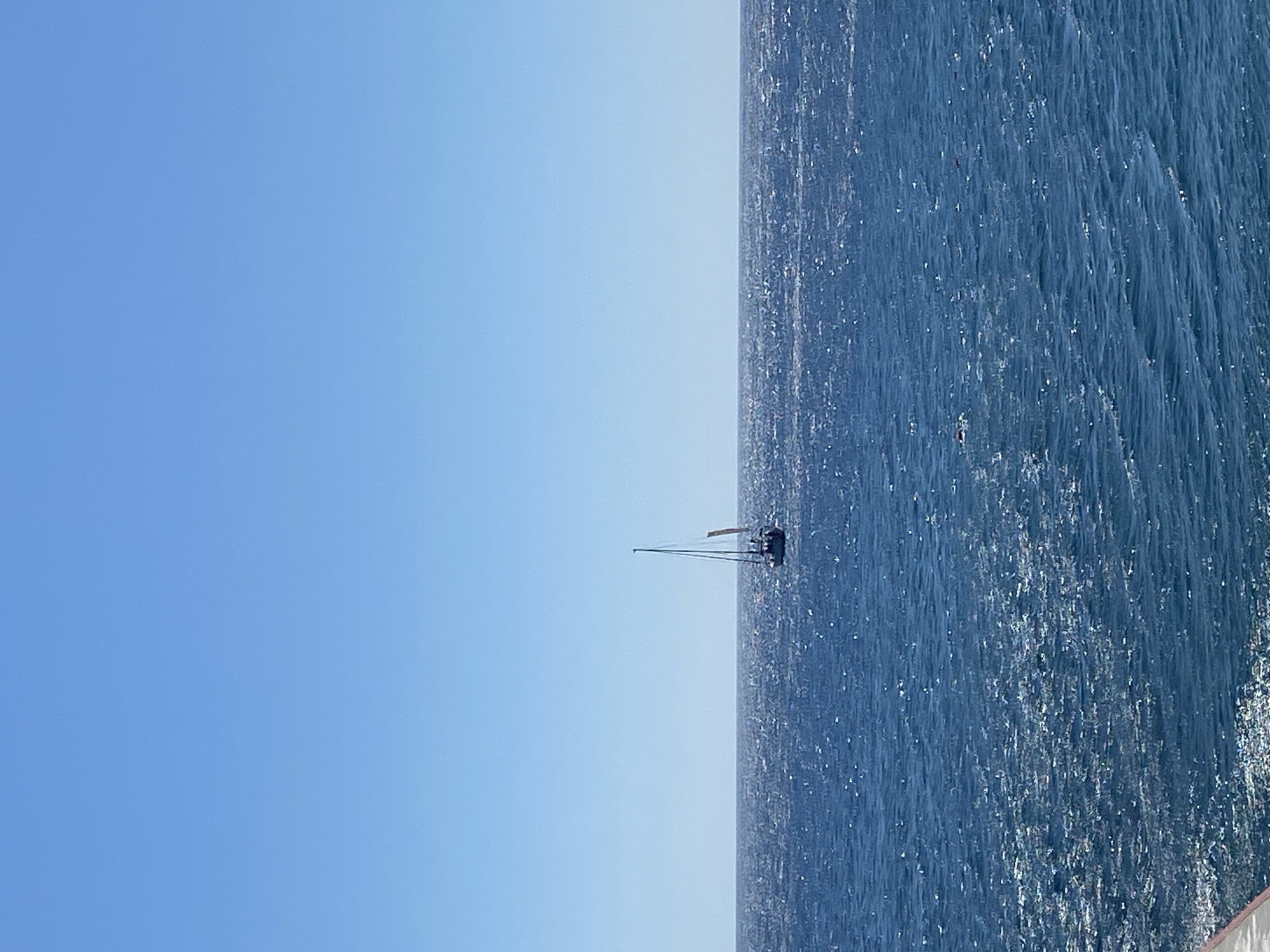 A sailboat with a single mast and sail is visible on the horizon in the middle of a vast expanse of deep blue ocean. The water is slightly choppy, with sunlight glinting off the surface. The sky above is a clear, bright blue. There appears to be a landmass or distant coastline on the far horizon. The image is taken from a slightly elevated perspective, possibly from a shore or higher vantage point.