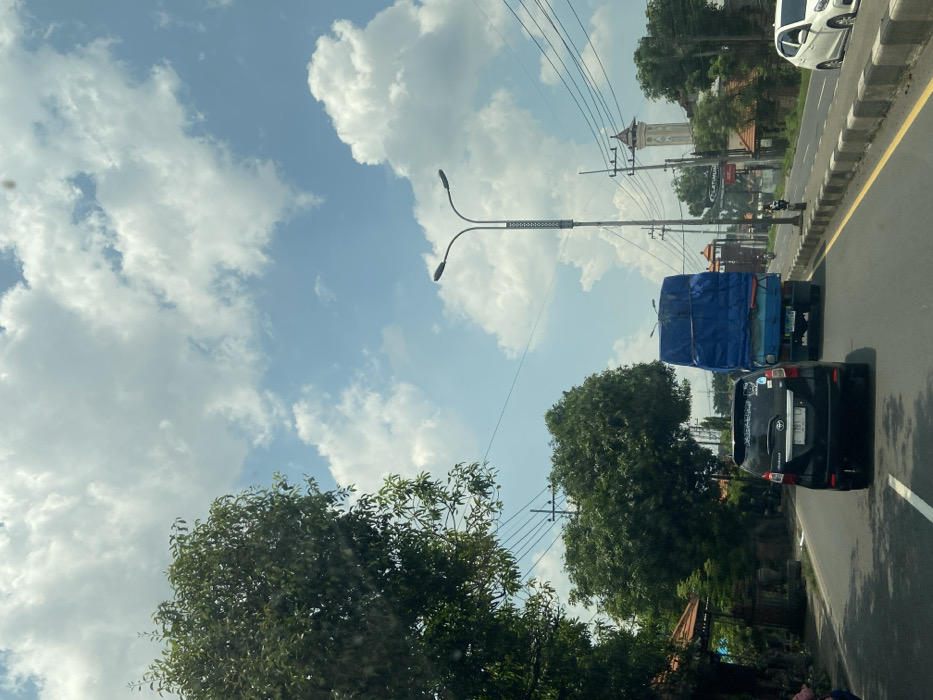 A daytime street scene shows moving traffic, including a black SUV and a blue-covered truck. Lush green trees flank the roadside, contrasting with a bright sky filled with scattered white clouds. Overhead power lines stretch across the scene, joining a modern street lamp. A tall, slender building with a distinct architectural style is visible in the distance, adding a touch of cultural interest. The photo is taken from a low angle, suggesting the perspective of a passenger in a vehicle.