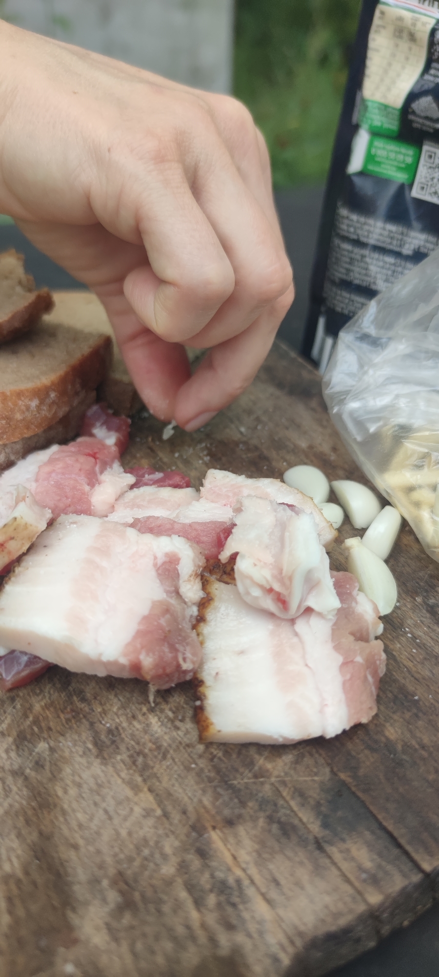 A person's hand is shown reaching down to pick up a tiny piece of food from a wooden cutting board. The board is covered with slices of cured pork belly, pieces of dark bread, and several cloves of garlic. A black bag with white lettering and a clear plastic bag are visible in the background. The overall scene appears to be set outdoors, possibly in a garden or on a patio, with blurred green foliage in the background. The lighting suggests it is daytime.