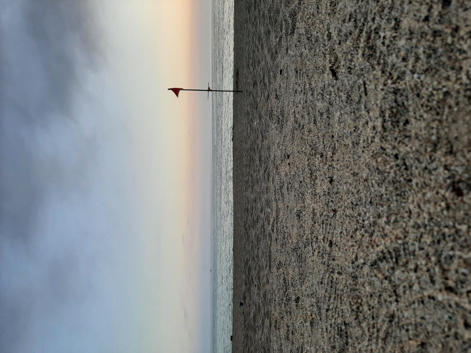 The image shows a beach scene, likely in Alanya, Turkey, given the provided context. The composition is divided roughly in half, with the right side showing coarse, dark sand and the left showing the ocean and sky. A red flag on a pole is positioned at the edge where the sand meets the water. The sky appears overcast with a gradient of blue-gray transitioning to a lighter, peach-toned horizon. The water is calm, with gentle ripples. There are no visible people or other distinct objects in the water or on the sand.