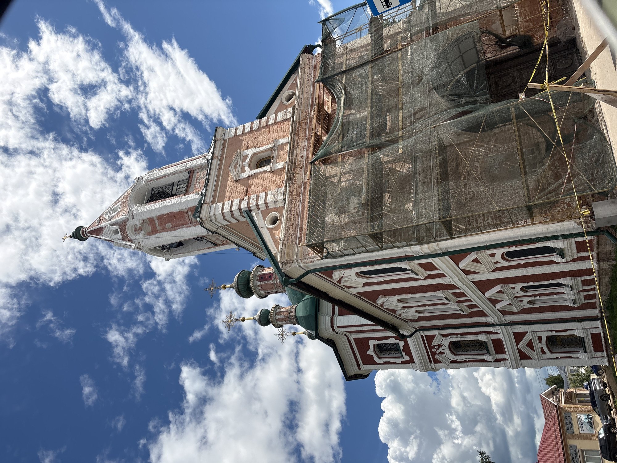 The image depicts the Church of John the Warrior in Pereslavl-Zalessky, Russia, on a bright, cloudy day. The church is an Orthodox Christian structure with red and white facades, multiple green domes topped with crosses, and a prominent bell tower. The bell tower is partially covered in scaffolding and netting, suggesting ongoing renovations. In the foreground, construction barriers and caution tape are visible. The background shows a clear blue sky with scattered white clouds and a few trees. In the lower left corner, a building with signs in Russian and parked cars can be seen.