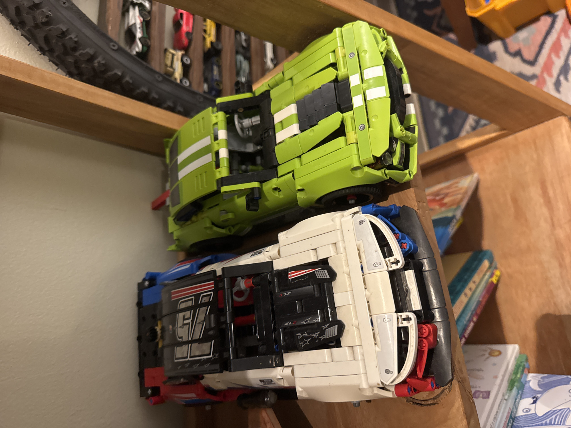 The image shows two Lego Technic car models stacked on a wooden shelf. The top model is green with white stripes. The bottom model is white with blue and red accents, and has the number 97 on its side. A black bicycle tire is mounted on the shelf above the green car. Several toy cars are visible in the background, along with books and a portion of a rug. The location is not discernible from the provided image. Therefore, the location cannot be stated.