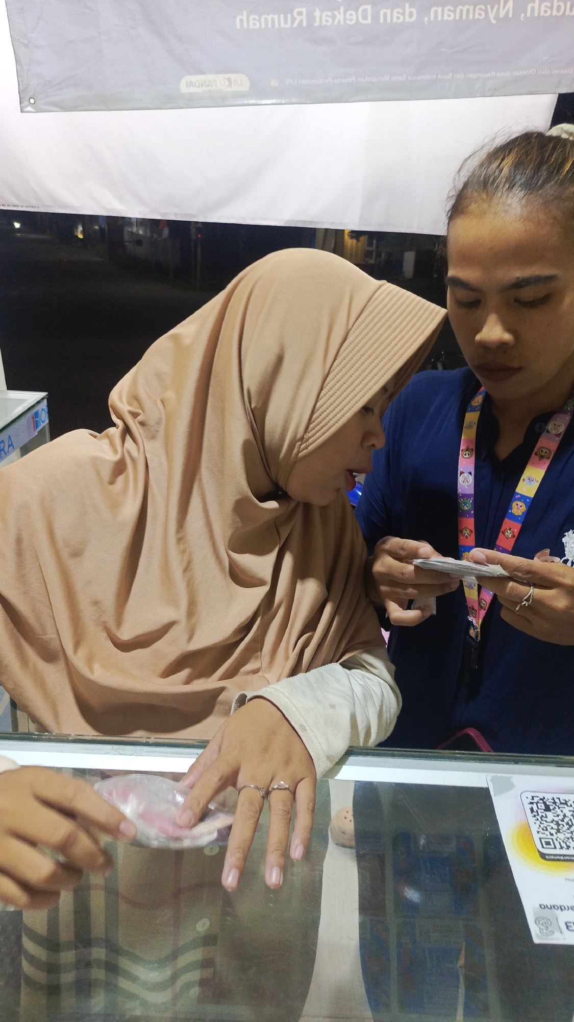 Two individuals are shown at a counter. The person on the left wears a tan hijab and a light-colored long-sleeved shirt. This individual is holding a stack of bills. The person on the right wears a dark blue shirt and has a lanyard with colorful charms around their neck. This individual is looking at the bills held by the person on the left, and their hands are near the bills. The counter appears to be made of glass, and reflections of the people and their surroundings are visible. In the background, a banner is displayed with text that is partially obscured and in reverse. A QR code is visible on the right side of the image. The scene appears to be outdoors at night, possibly in Parung Panjang, Indonesia, given the banner's likely reference to a local business or event.
