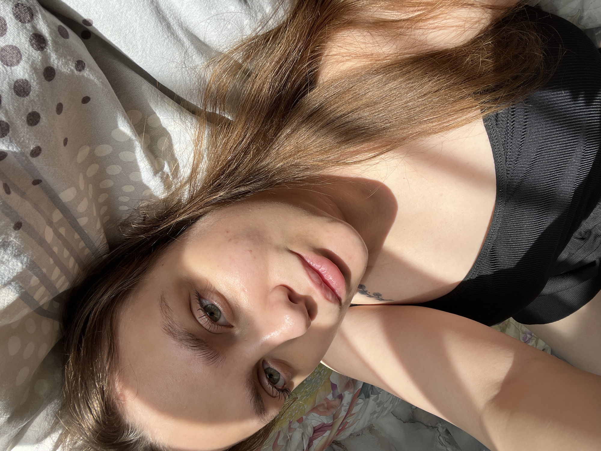 A woman with long, light brown hair is lying on a bed. She is wearing a black top. Her face is partially lit by sunlight, casting shadows. The bedsheets have a gray and white polka dot pattern and a floral pattern. A tattoo is visible on her upper chest.