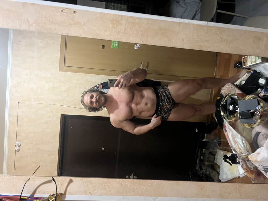 A muscular man poses in front of a mirror, wearing patterned underwear. His right arm is flexed. Behind him are a beige wall and a dark door, and there's a mess of items on the floor to his right. The lighting is soft and warm. The image is taken from a slightly distorted perspective due to the mirror's angle.