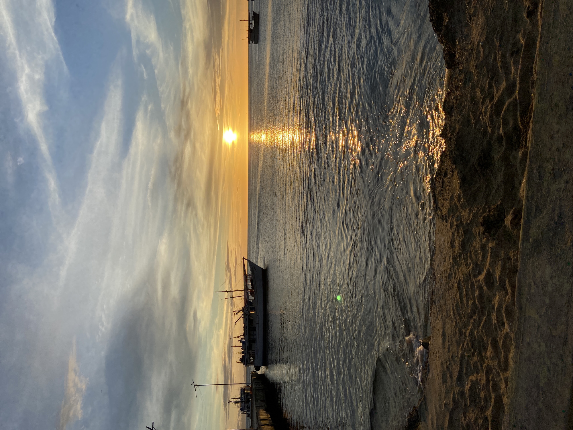 This image captures a sunset over the sea in Juan Griego, Venezuela. The sky is filled with a mix of blue and white clouds, with streaks of orange and yellow near the horizon where the sun is setting. The sun itself is a bright, glowing orb, casting a shimmering reflection on the water's surface.
In the foreground, a sandy beach meets the calm sea. Some rocks are visible at the edge of the water. A concrete walkway or pier extends into the water on the left side of the image.
Several boats are present in the scene. A large, white boat is docked at the pier, appearing to be a fishing vessel with multiple masts and rigging. In the distance, another smaller boat is visible on the right side of the frame, also silhouetted against the sunset. A third boat is partially visible behind the pier on the left.
There are no people clearly visible in the image. The overall atmosphere is serene and peaceful, with the natural beauty of the sunset and the calm sea dominating the scene. The presence of the boats suggests a maritime community or port activity.