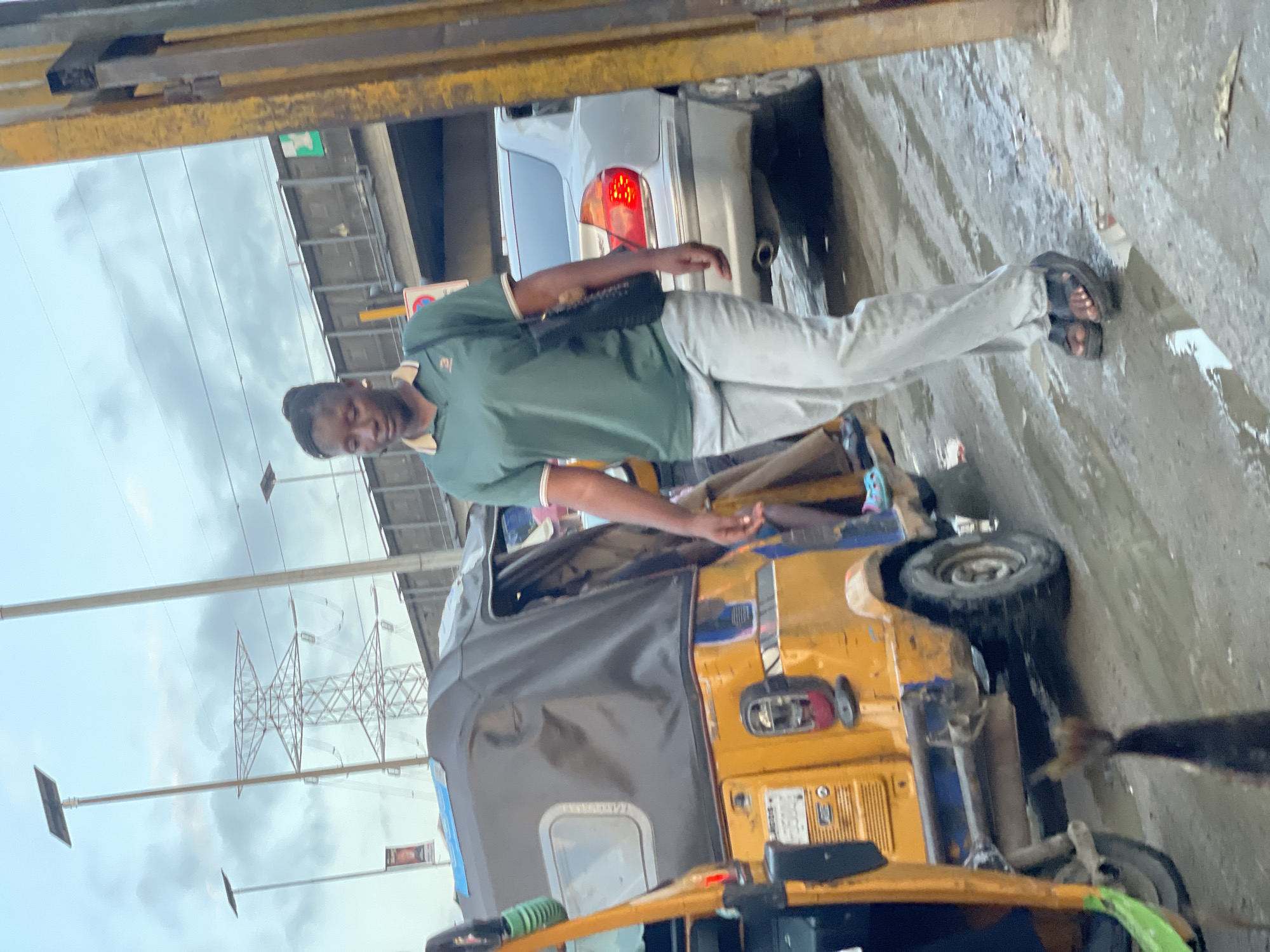 The image depicts a street scene in Lagos, Nigeria, likely during the late afternoon or early evening, given the overcast sky. A young woman, wearing a green polo shirt and light-colored trousers, is walking towards the right of the frame. She carries a black handbag over her left shoulder. To her left, a yellow and gray auto-rickshaw, a common mode of transport in Lagos, is parked. Its door is ajar, revealing a glimpse of its interior. In the background, a silver sedan with its brake lights illuminated is visible. The ground is wet and appears to be a road or street, with puddles reflecting the subdued light. Overhead, power lines and a large electricity pylon can be seen against the cloudy sky. The overall impression is one of bustling urban life, typical of Lagos.