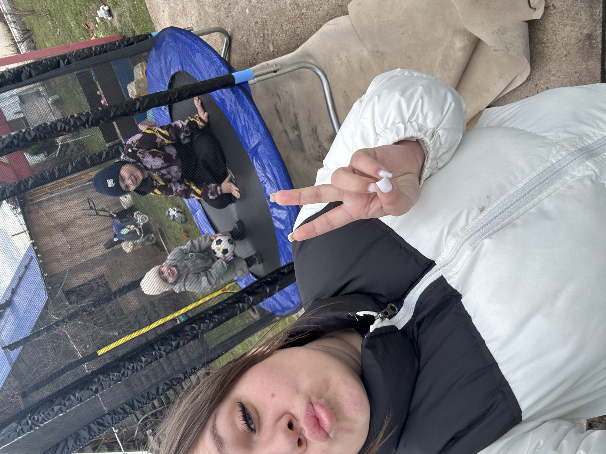 The image appears to be a selfie taken outdoors. In the background, there is a trampoline with two children on it. One child is lying down, and the other is sitting and holding a soccer ball. The surrounding environment includes grass, a building, and a patch of concrete. The location is Ольховка, Russia. The primary subject of the photo is the person taking the selfie, who is making a peace sign.