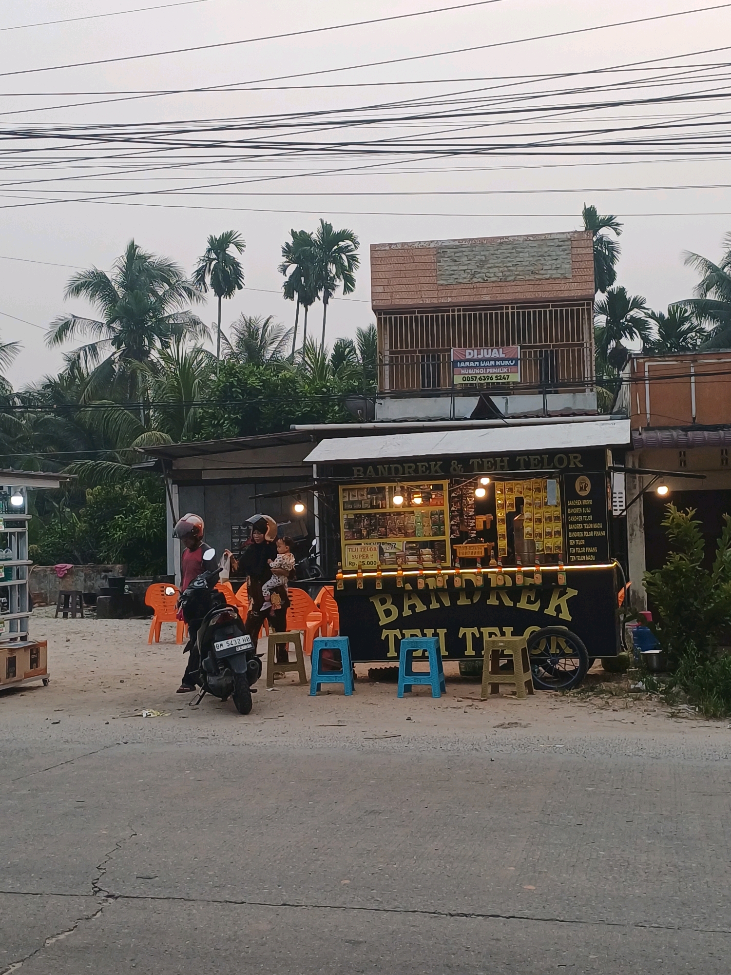 The image depicts a street scene in Dumai City, Indonesia. In the foreground, a paved road with visible cracks runs horizontally across the frame. To the right of the road, a small food cart labeled "BANDREK & TEH TELOR" is prominently displayed, featuring illuminated signage and various products for sale. Several orange plastic chairs are arranged in front of the cart, with two children's chairs, one blue and one yellow, also visible. A motorcycle with license plate "BM 5432 HB" is parked to the left of the food cart, and a person wearing a helmet is seated on it. Behind the motorcycle, a woman holding a child stands near a person in a red shirt also wearing a helmet. In the mid-ground, several structures with covered roofs are visible, with one building in the background featuring a sign that reads "DIJUAL" (For Sale). A dense backdrop of palm trees and foliage frames the scene. The sky is overcast with a network of power lines crisscrossing the upper portion of the image.