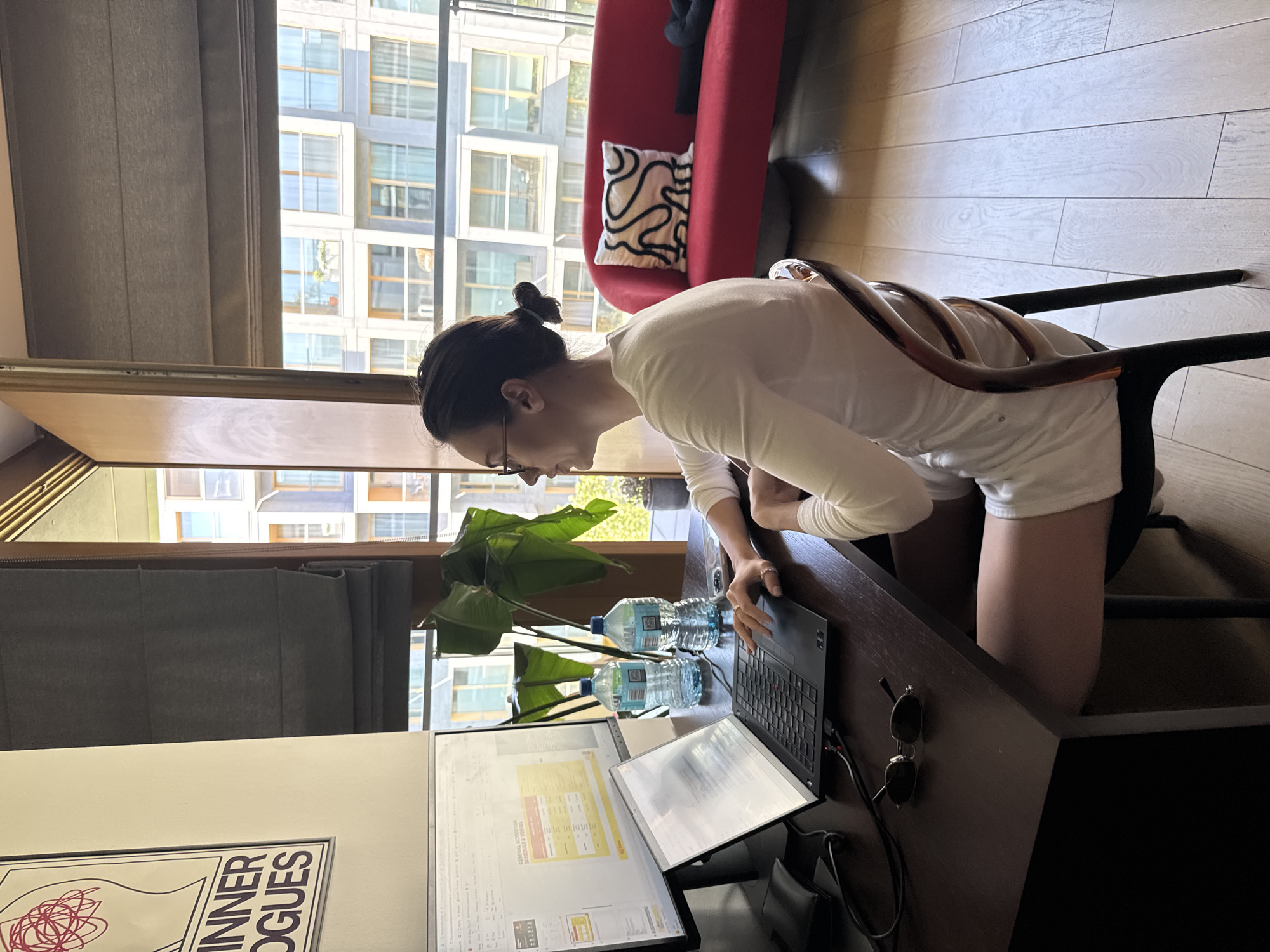 A woman is seated at a desk in an indoor setting, likely an apartment or office space, in the city of Unknown, Unknown. She is wearing a white long-sleeved shirt and shorts. She is positioned in front of a laptop computer and two additional monitors, appearing to be working. Two water bottles are placed on the desk beside the laptop. Sunglasses are also on the desk, along with a black bag. A houseplant sits between the woman and the monitors. A red sofa with a patterned pillow is visible in the background. Outside the window are apartment buildings. The floor is light-colored wood. A poster is leaning against the wall. A gray window shade is pulled down.
