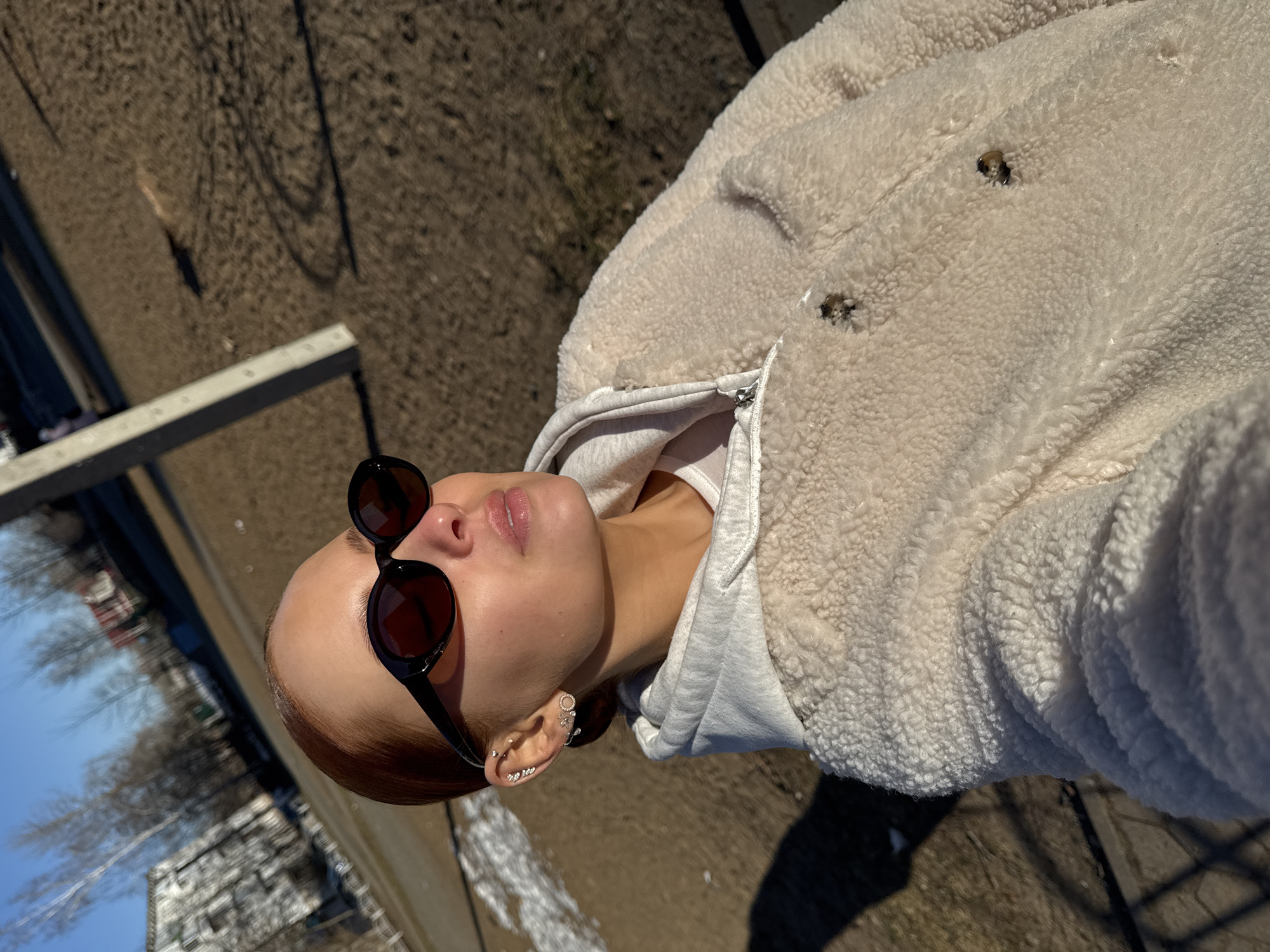 A woman with fair skin and auburn hair pulled back is looking upwards while wearing sunglasses, a white hoodie, and a light-colored fleece jacket. She has multiple earrings in her left ear. The photo appears to be taken outdoors on a sunny day. The background includes a blurred view of buildings and bare trees, suggesting a street scene. Patches of snow are visible on the ground. The photo might be taken in the city of Chaikovsky, Russia, as the request includes this location, though this can't be explicitly confirmed from the image.