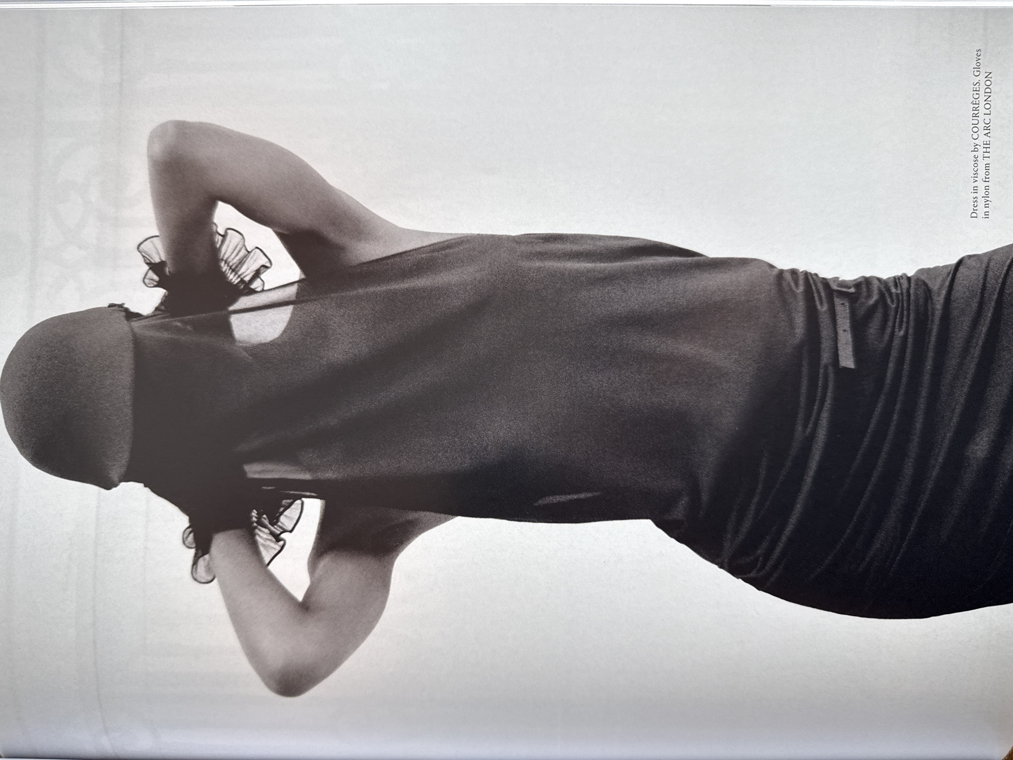 A black and white image shows a woman in a Courrèges viscose dress and Courrèges nylon gloves from The Arc London. The woman is positioned with her back to the viewer and her head slightly turned to the left. Her arms are raised and bent with her hands near her head. She appears to be wearing a cap or hood that covers her hair. The background is a plain white. The image is from a magazine.