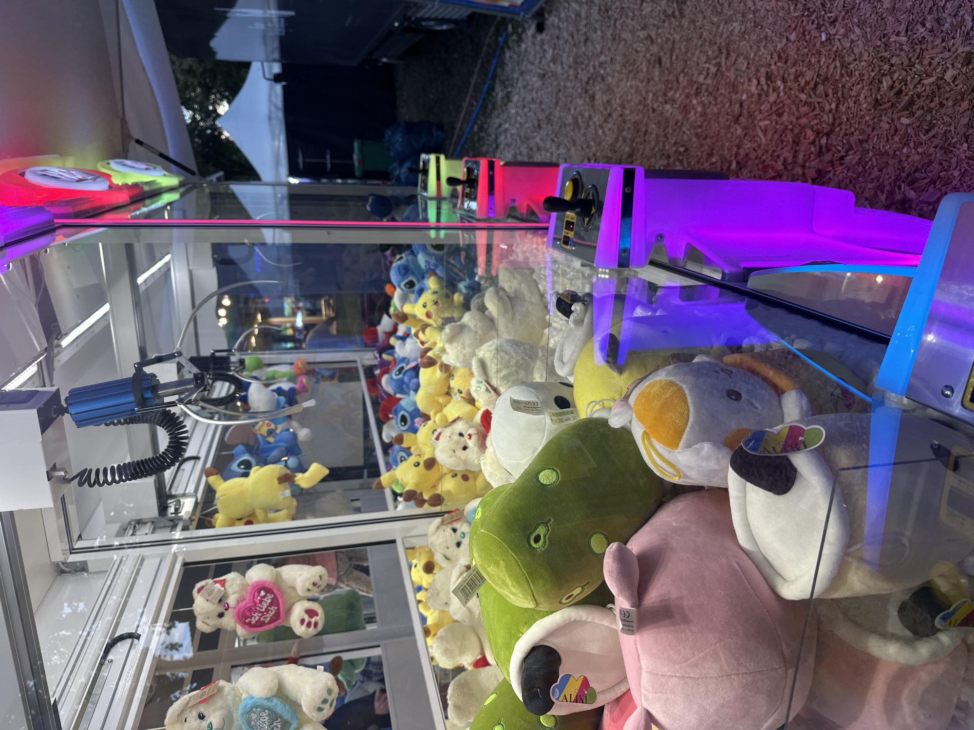 The image shows a claw machine filled with plush toys, situated outdoors at the "Bruchhausen-Vilsen Summer Festival" in Bruchhausen-Vilsen, Germany. The claw machine has a clear glass front and is illuminated with purple and pink neon lights around its controls. Inside, a variety of stuffed animals are visible, including teddy bears, yellow Pikachu-like characters, blue Stitch characters, and green avocado-shaped pillows. In the background, a white tent and a dark-colored truck are partially visible, suggesting a festival or fairground environment. The ground is covered with wood chips. No people are clearly visible interacting with the machine in this specific frame.