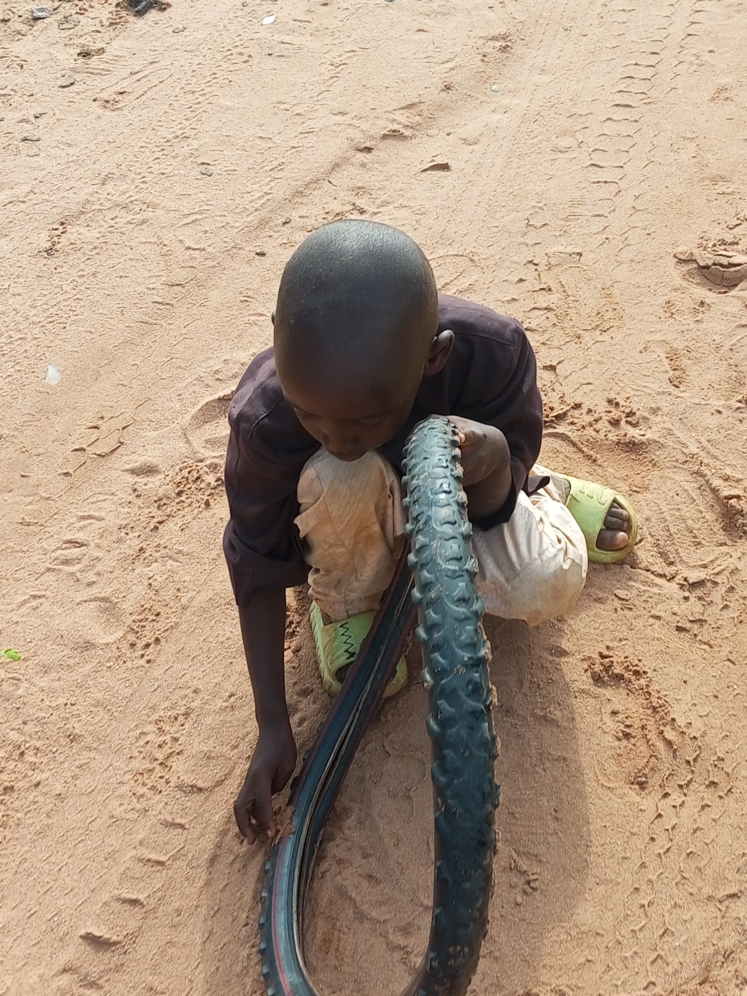 A dark-skinned child is kneeling on sandy ground in Tempure, Nigeria. The child is wearing a dark shirt and light-colored pants, with bright green sandals on their feet. They are holding a tire, which appears to be part of a bicycle, and leaning over it, with their head close to the tire. The ground is predominantly sand, with some tire tracks visible in the background. There are also small indentations in the sand, possibly from footprints or other objects. The lighting suggests it is daytime.