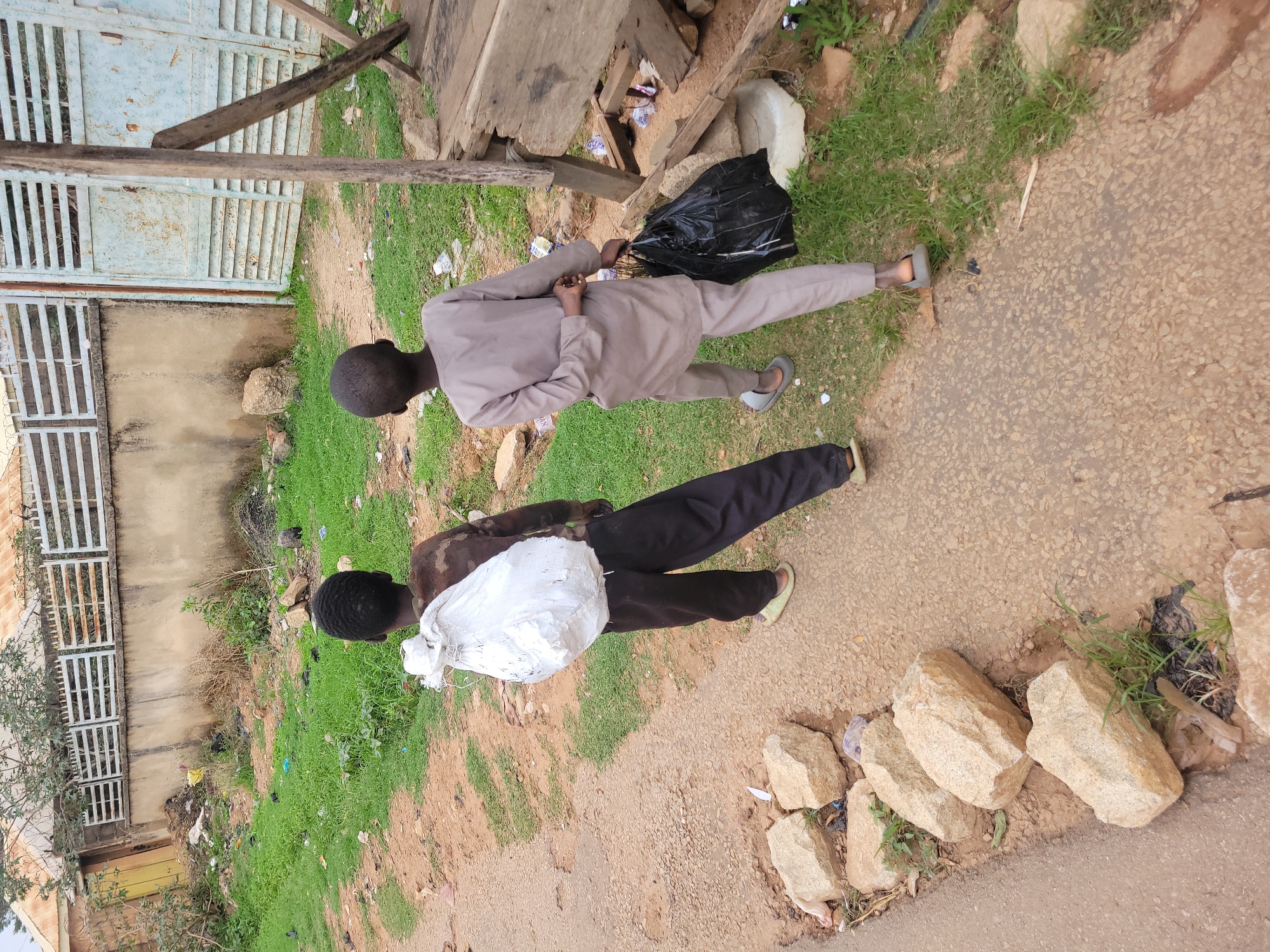 Two young men are walking along a dirt path in Jos, Nigeria. One man is carrying a large white bag, while the other is carrying a black trash bag. The path is lined with grass, rocks, and trash. A weathered gate can be seen in the background.