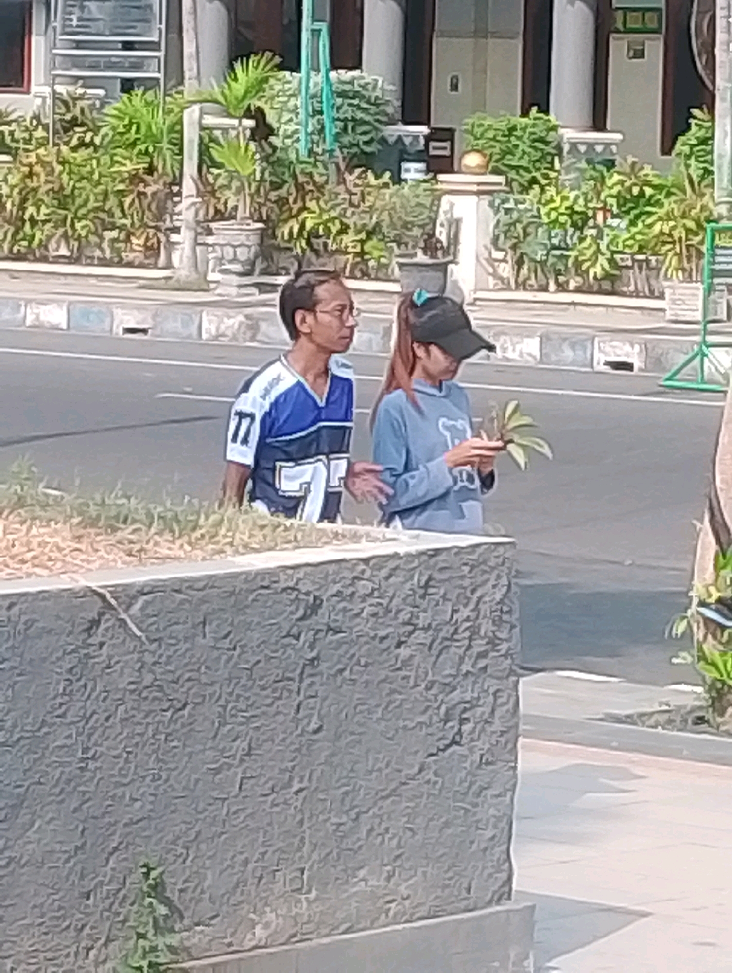 The image shows two individuals on a street in Probolinggo, Indonesia. On the left, a man wearing glasses and a blue and white jersey with the number 77 is facing right. To his right, a young woman wearing a black baseball cap and a blue hoodie is holding a small plant in her hands. They appear to be standing on the side of a paved road, with a textured stone wall in the foreground and a sidewalk and buildings with greenery in the background. The lighting suggests it is daytime.