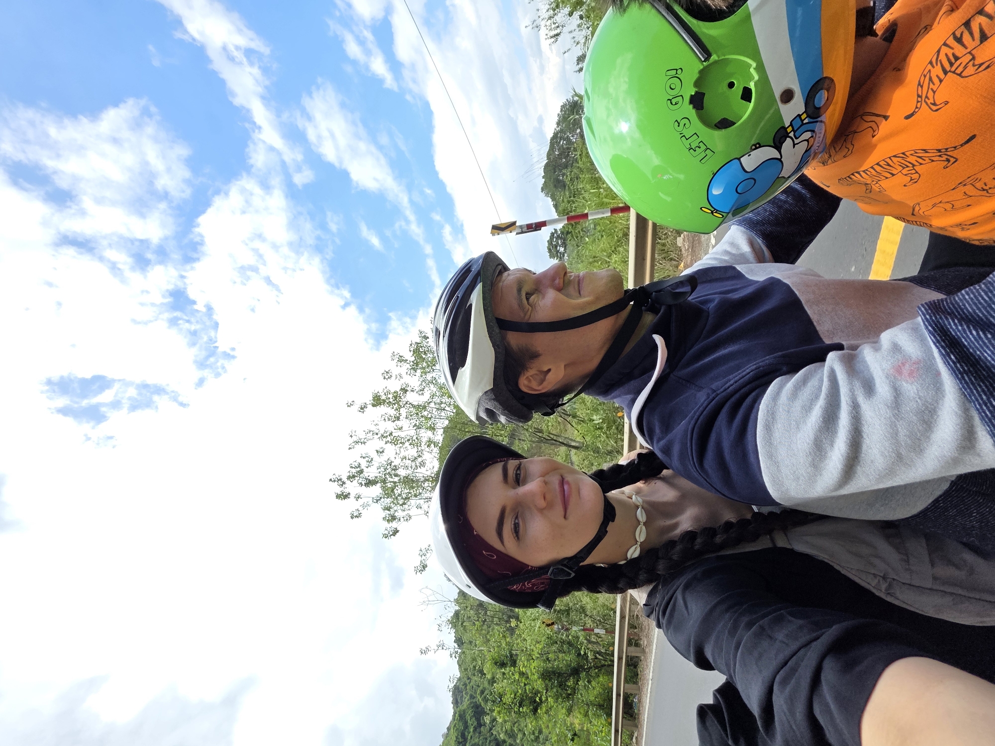Three people are captured in a selfie. The setting appears to be Thôn 5, Vietnam, as indicated by the mountainous landscape and vegetation.
From front to back:
- A woman wearing a white helmet with a burgundy bandana beneath it, a black hoodie, and a seashell necklace. Her hair is braided.
- A man with a white and black helmet. He is wearing a navy and gray striped sweatshirt.
- A child wearing a green helmet with "Let's Go!" written on it and an orange tiger-striped hoodie.
They are on a road with a yellow dividing line, a metal guardrail, and a sign with red and white stripes. The sky is blue with scattered clouds.