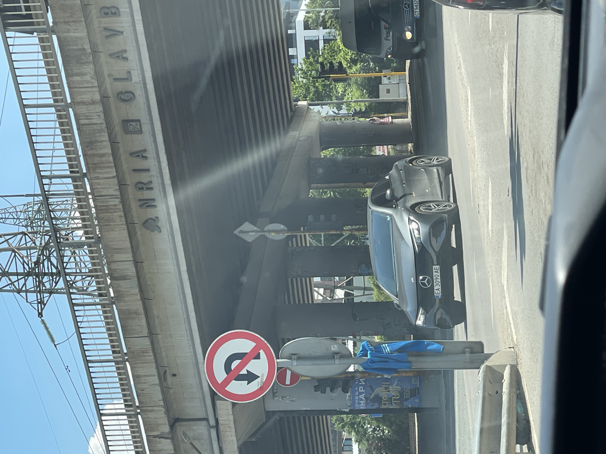 The image captures a street scene in Sofia, Bulgaria, dominated by an elevated road or bridge structure overhead. Underneath this overpass, traffic is moving along a road. A grey Mercedes-Benz EQE electric sedan with license plate "EA 3090 AE" is prominently visible in the foreground, traveling towards the viewer. To its right, a white Fedex van is also present. A "No U-turn" traffic sign is mounted on a pole on the left side of the road, accompanied by a smaller red and white "No Entry" sign. A blue garment, possibly a jacket or shirt, is hanging on the traffic sign pole. The bridge structure above is concrete, with what appears to be lettering or symbols embossed on its underside. Tall power lines and pylons are visible against a clear blue sky, indicating an urban or suburban setting. Green trees and buildings can be seen in the background, suggesting a daytime setting. The image is taken from within a vehicle, as evidenced by the dashboard or interior elements visible at the bottom of the frame.