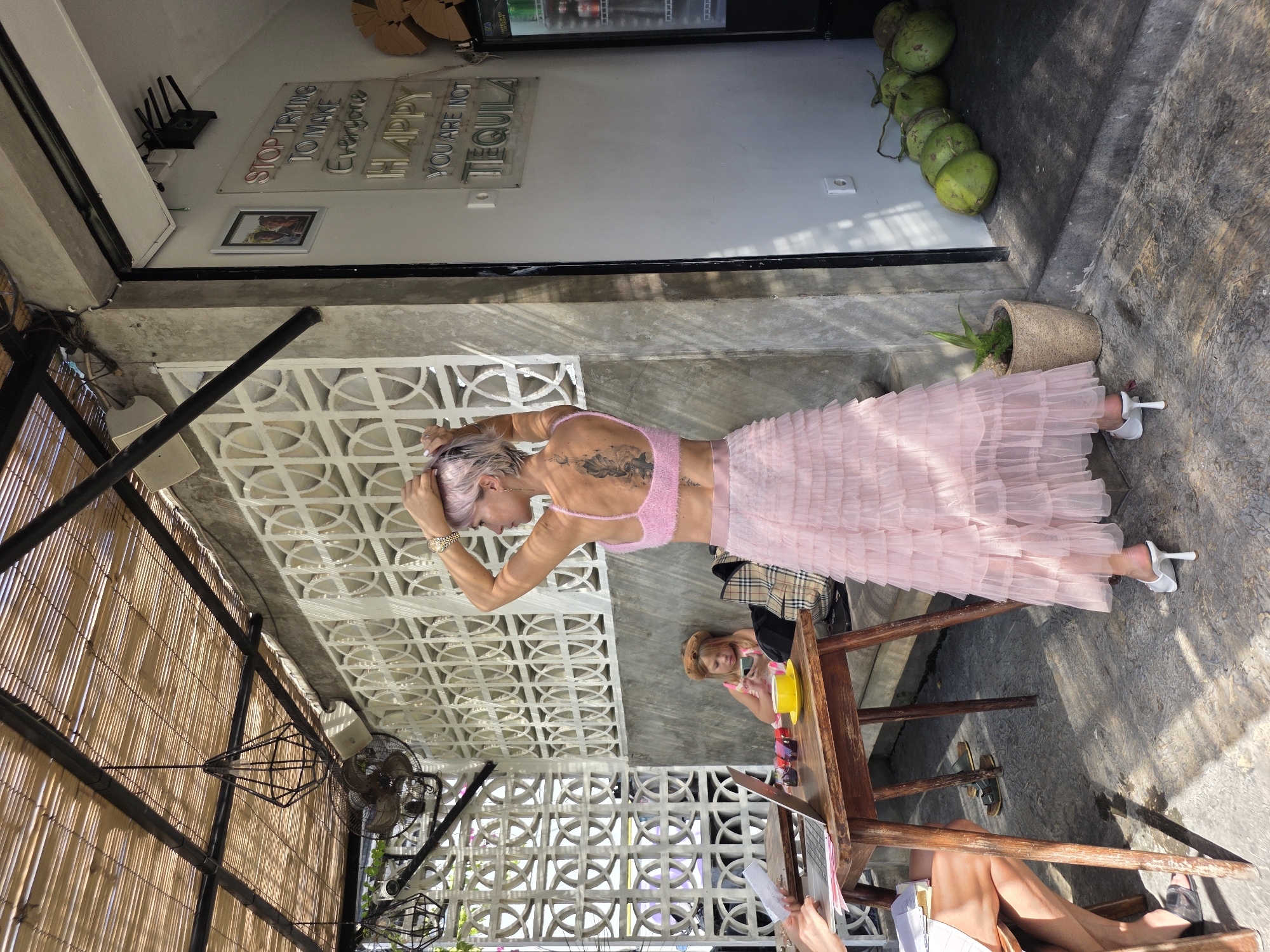 The image depicts an outdoor cafe area in North Kuta, Indonesia. A woman with short pink hair and a large back tattoo is standing, facing away from the camera, with her hands on her head. She is wearing a pink lace bralette and a long, pink tiered tulle skirt, along with white heeled mules. In the background, a young girl with blonde hair is seated at a wooden table, looking at her phone. Another person's legs are visible on the left side of the frame, near a laptop and papers on the table. The cafe has a bamboo-covered ceiling, a decorative white wall, and concrete elements. A neon sign on the wall reads, "STOP TRYING TO MAKE EVERYONE HAPPY YOU ARE NOT TEQUILA". A row of green coconuts is lined up near the entrance of the cafe.
