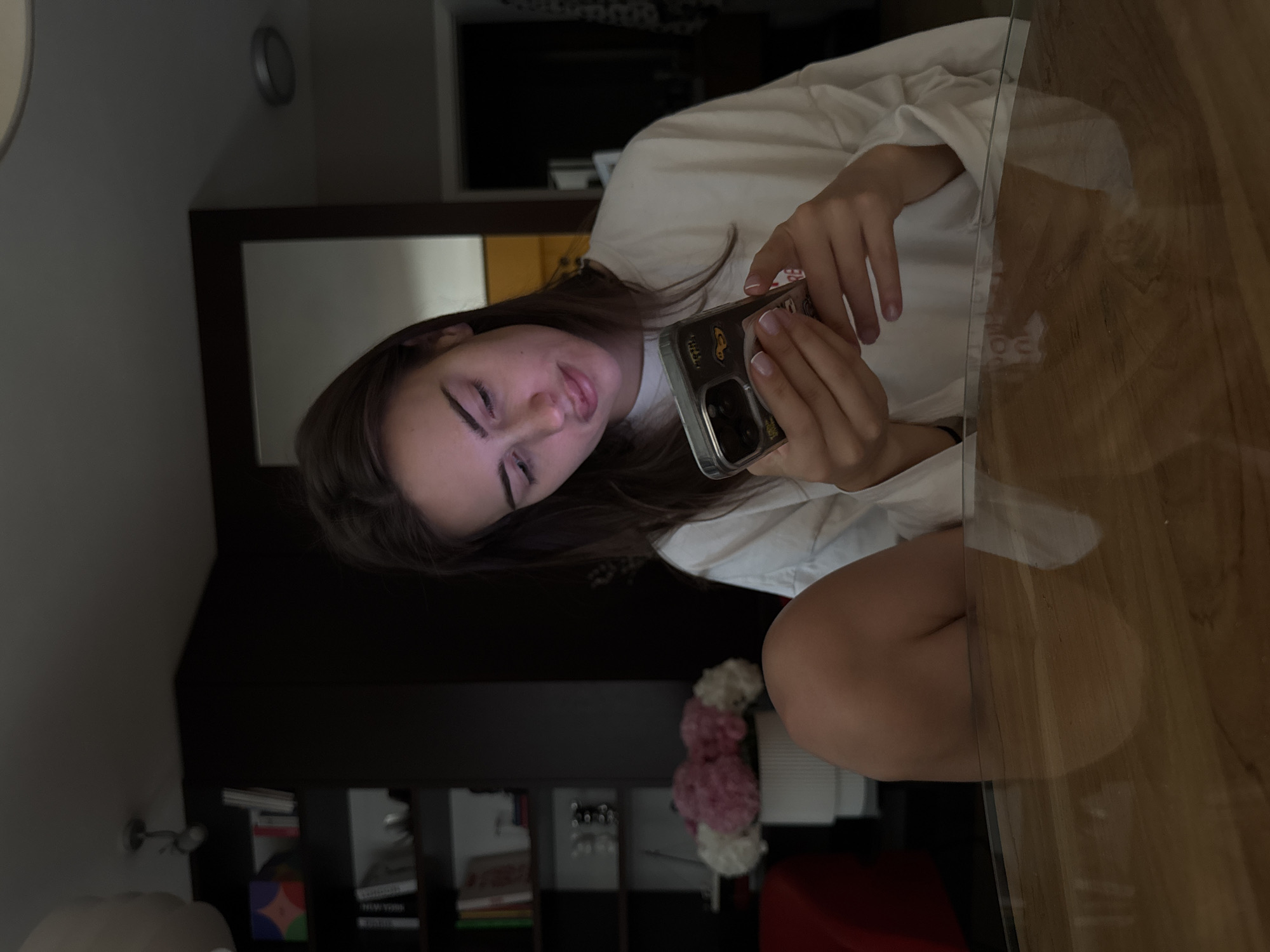 A young woman sits at a clear-topped table with a wood-grain base. She is looking down and holding a cell phone in her hands. She is wearing a white long-sleeved garment. The background contains a dark-colored bookcase with visible books and a mirror. A clock is mounted on the wall. A bouquet of flowers is visible near the base of the bookcase. It is unknown if this photo was taken in Warsaw, Poland.