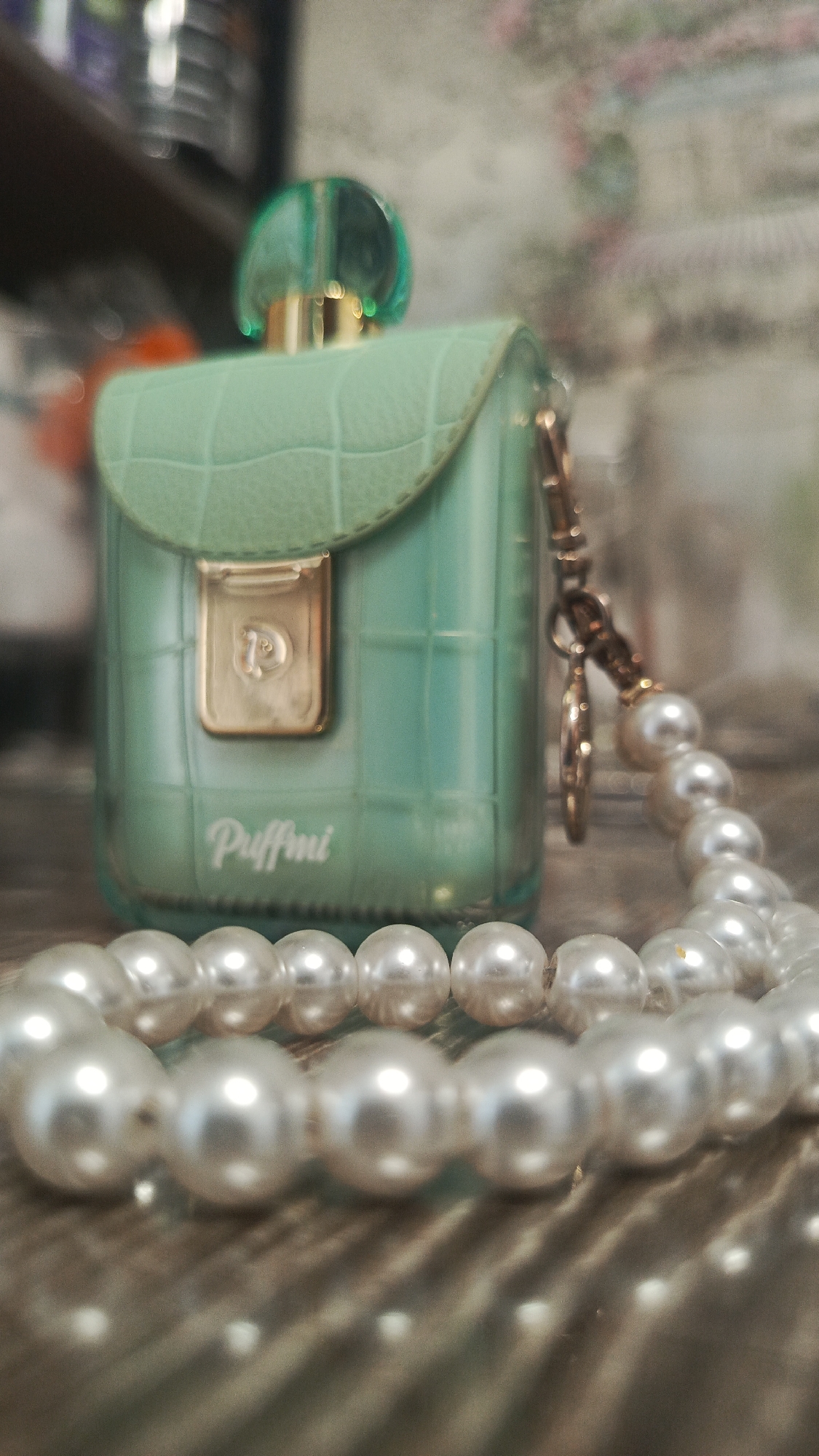 A green, purse-shaped perfume bottle with a textured lid sits on a wooden surface, partially obscured by a strand of pearl beads in the foreground. The bottle has a gold clasp with a "P" logo and the word "Puffmi" printed below it. A gold chain is attached to the side of the bottle, with a small charm hanging from it, also partially covered by pearls. The background is blurred and appears to be an interior setting with shelves and objects. There is no indication that this scene is located in Zvenigorod, Russia.