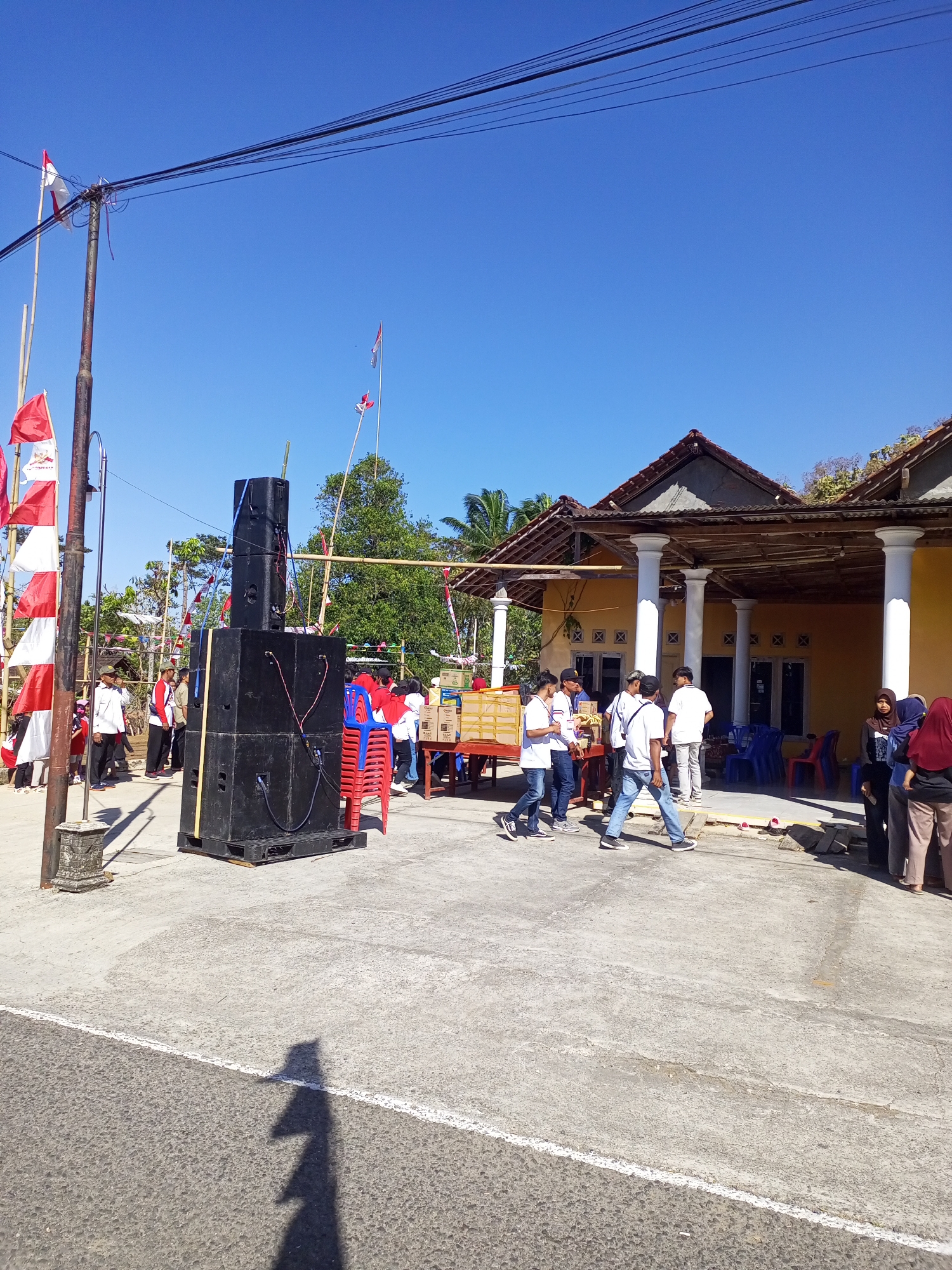 An outdoor scene in Jatipurno, Indonesia, depicts a gathering with several people milling about. In the foreground, large black speakers are stacked on a pallet. To the left, flags are mounted on poles, including red and white flags. Behind the speakers, a table is set up with boxes, likely for distribution or display. Red and blue chairs are stacked nearby. In the background, a yellow building with a tiled roof and columns stands under a clear blue sky. A group of men, some wearing white shirts, are gathered near the table. Women are also present, some wearing headscarves. The ground is paved with concrete and asphalt.