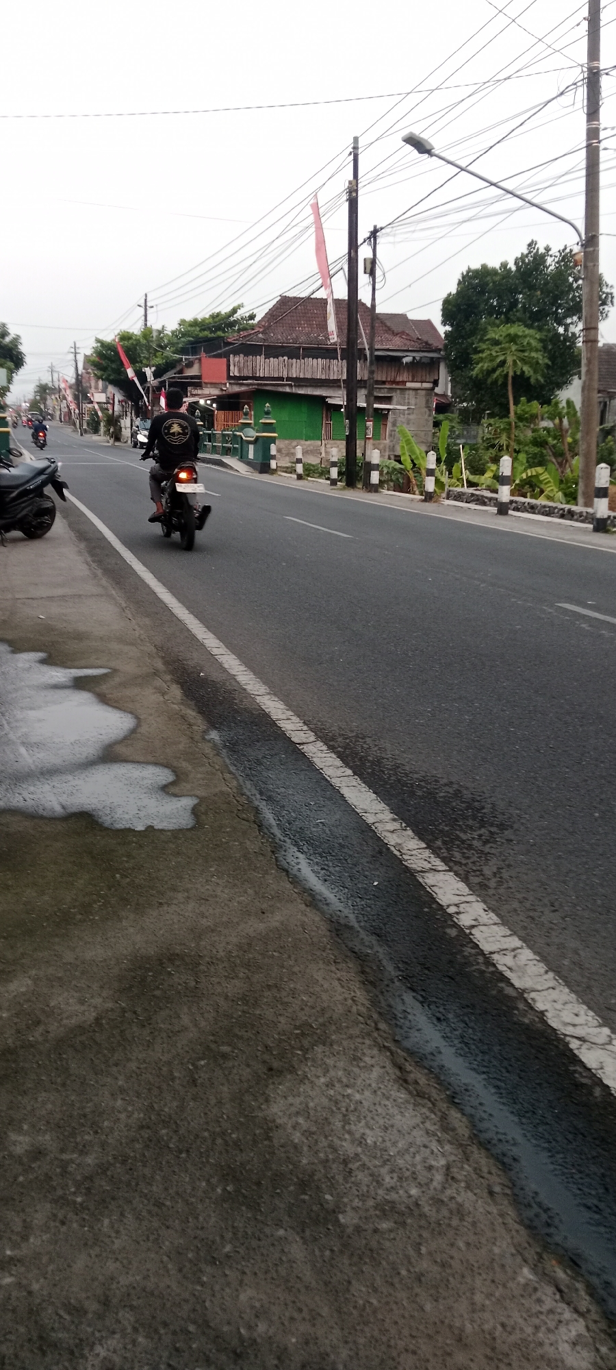 A motorcyclist, wearing a black shirt with a graphic and grey pants, is riding a black motorcycle on a wet, grey asphalt road. The motorcycle has a white license plate illuminated. The road has a white broken line marking the lane. The scene is set in Maguwoharjo, Indonesia, suggested by the presence of Indonesian flags (red and white) mounted on poles along the roadside. Buildings with red tiled roofs and green painted walls are visible in the background. Several power lines are suspended overhead, supported by poles. The sky is overcast, indicating a cloudy day. The foreground shows a wet concrete sidewalk with several puddles of water. Other parked motorcycles and a lamppost are also visible in the scene.