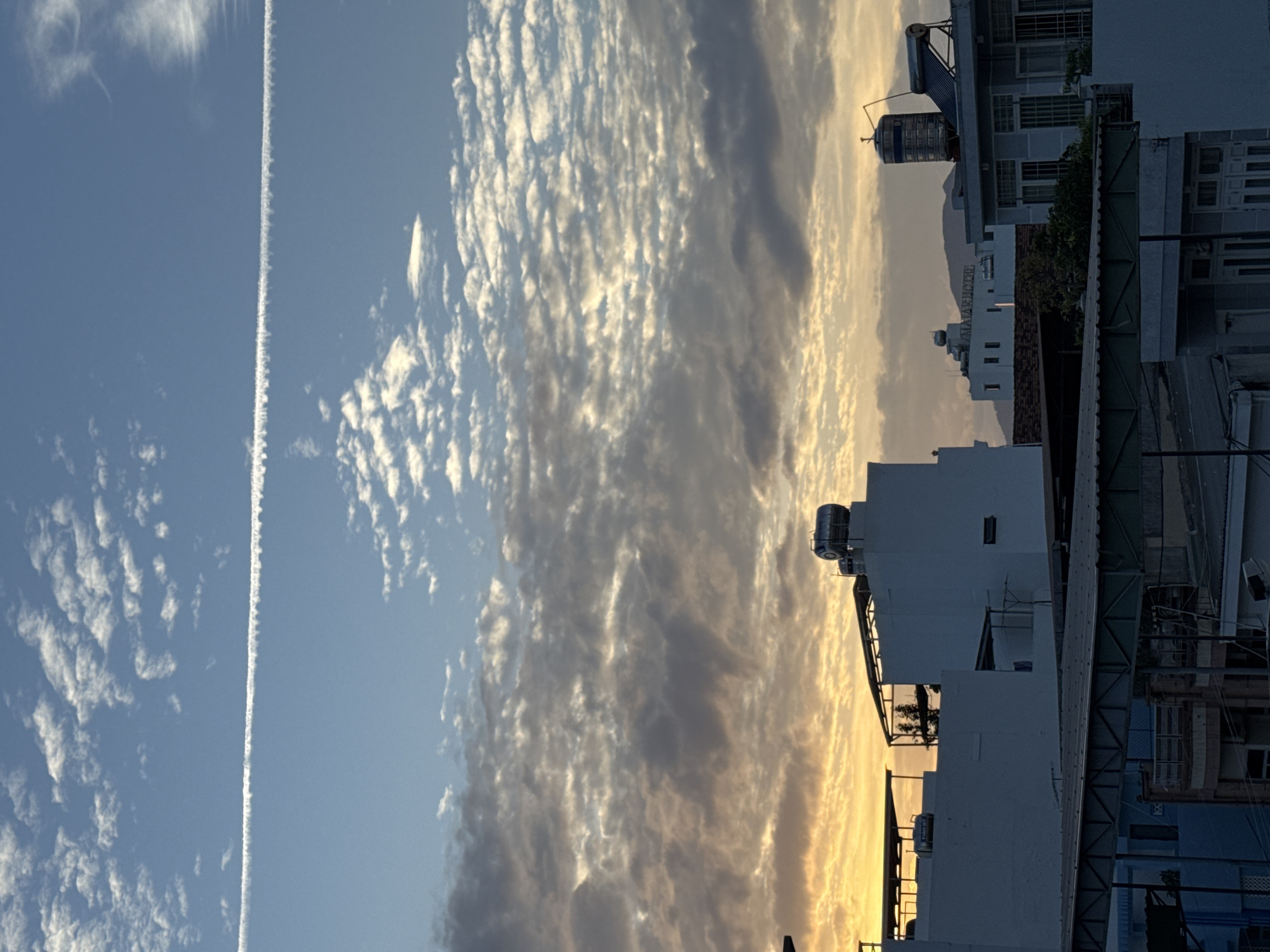 This is a photograph taken from a rooftop in Nha Trang, Vietnam, overlooking a cityscape at sunset. The sky is a gradient of blue and orange, with scattered clouds. A prominent contrail stretches horizontally across the upper portion of the image, indicating an airplane's passage. The foreground features a dense arrangement of rooftops and buildings, characterized by white facades, metal canopies, and water tanks. In the middle ground, several multi-story buildings are visible, some with signs such as "ALANI HOTEL". In the background, a mountain range is faintly visible under the hazy sky. The scene captures the urban landscape of Nha Trang bathed in the warm light of dusk.