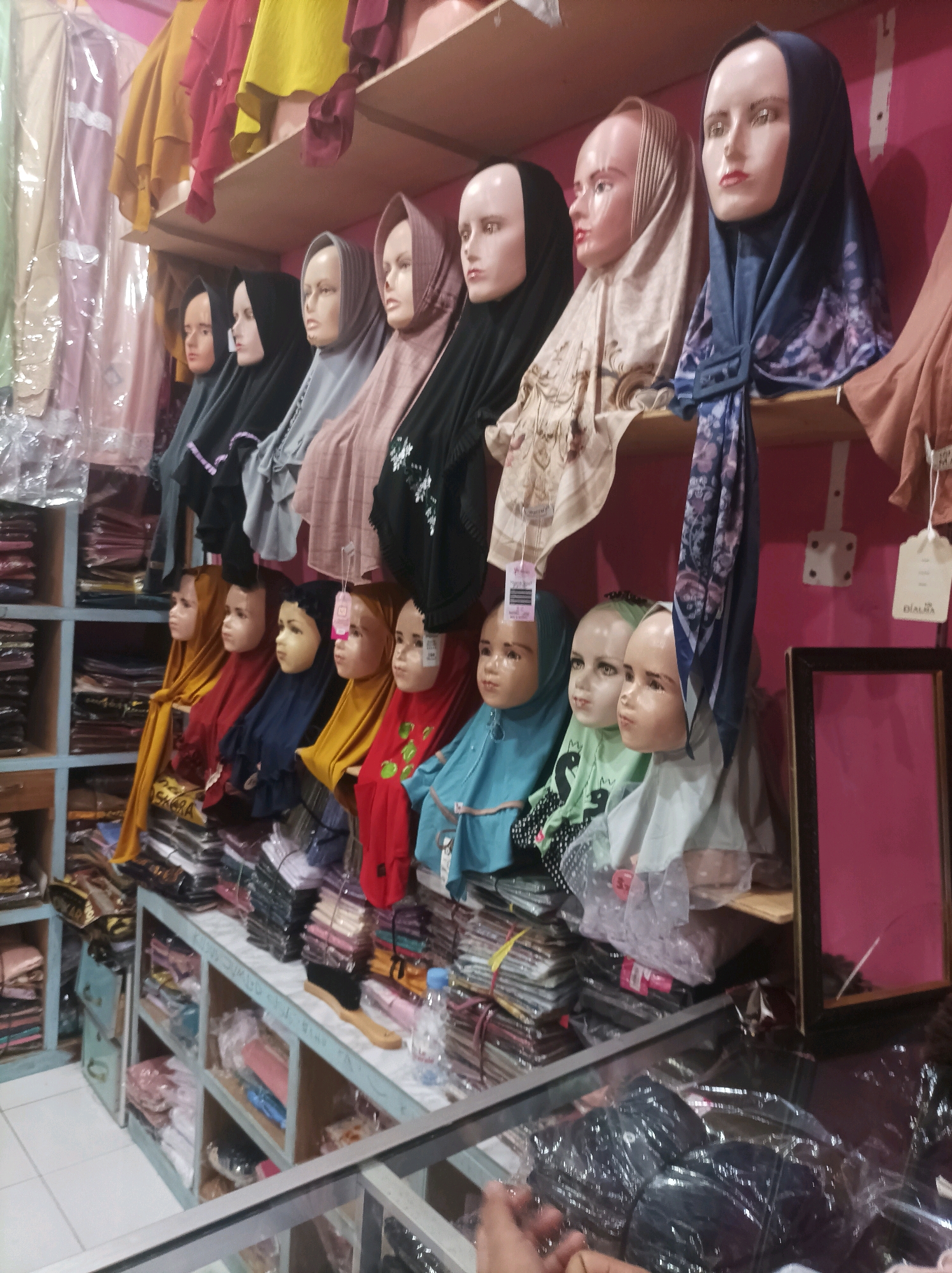 The image shows a retail store in Sumuragung, Indonesia, with shelves stacked with clothing, primarily hijabs and dresses, displayed on mannequins and folded on shelves. In the foreground, a counter with a glass display case is visible. The store appears to be well-stocked and organized. The lighting is bright and even, highlighting the colors and textures of the merchandise. The overall impression is of a bustling marketplace with a wide variety of clothing options.