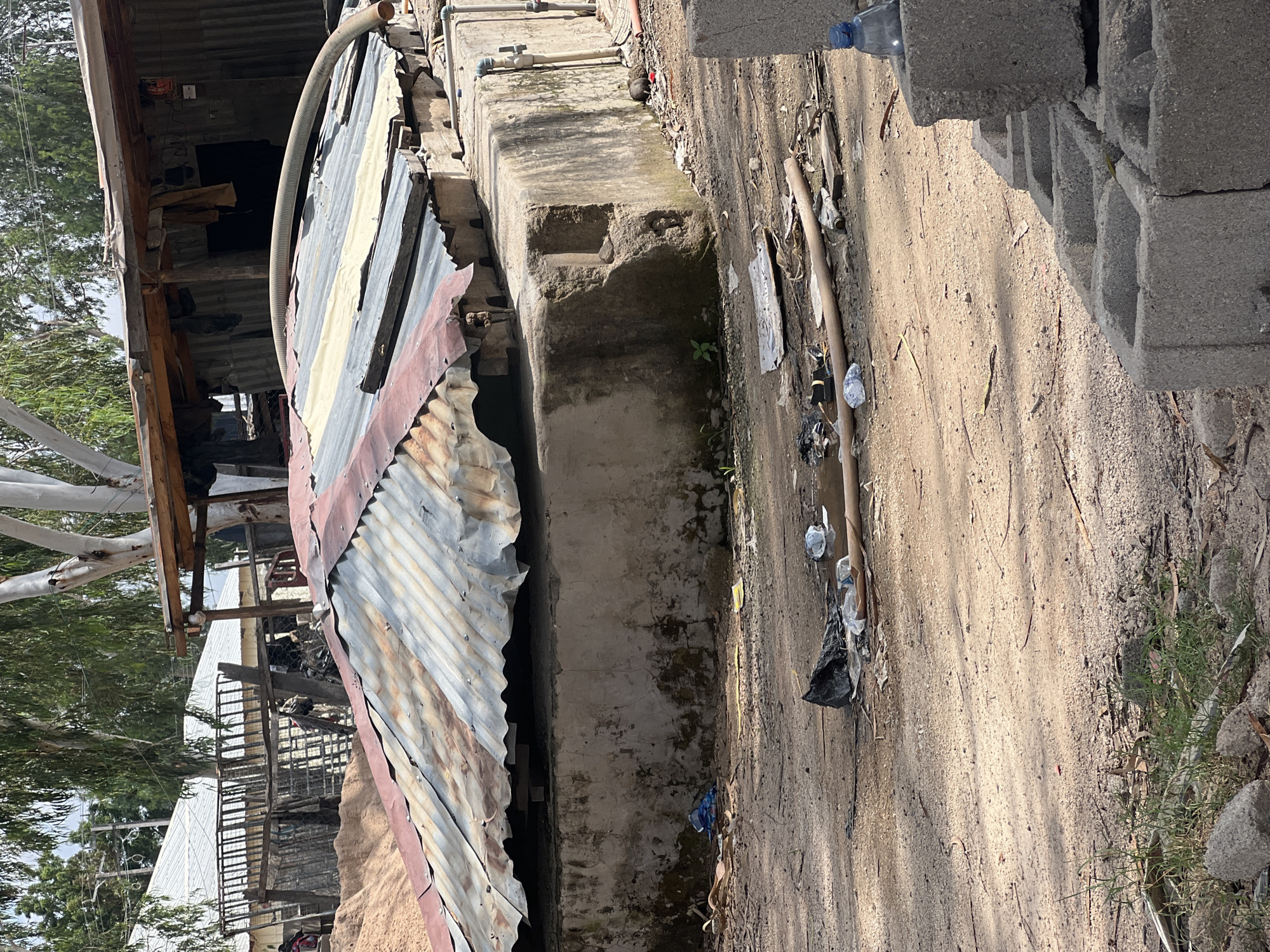 The image depicts an outdoor scene in Howayi, Nigeria, characterized by a rough, unpaved ground with sand and debris. A corrugated metal structure with weathered, multi-colored panels forms a prominent element on the left side of the frame, suggesting a makeshift shelter or building. Adjacent to this structure is a concrete ledge or platform, showing signs of age and possibly some vegetation growth.
The ground is littered with various objects, including plastic bottles, trash bags, and what appear to be discarded items. A wooden pole or stick lies horizontally on the ground. In the background, trees and foliage are visible, indicating a natural environment surrounding the constructed elements. To the right, a stack of grey concrete blocks is piled up, hinting at ongoing construction or material storage. A partially visible plastic bottle with a blue cap rests on top of these blocks.
The lighting suggests it is daytime, with shadows cast on the ground, indicating a sunny day. The overall impression is one of a utilitarian or possibly impoverished area, with elements of construction and outdoor living. No people are clearly visible, and the activity taking place is not overtly suggested, other than the presence of construction materials. The scene appears to be a factual representation of the environment without any overt emotional tone. No legible text is visible in the image.