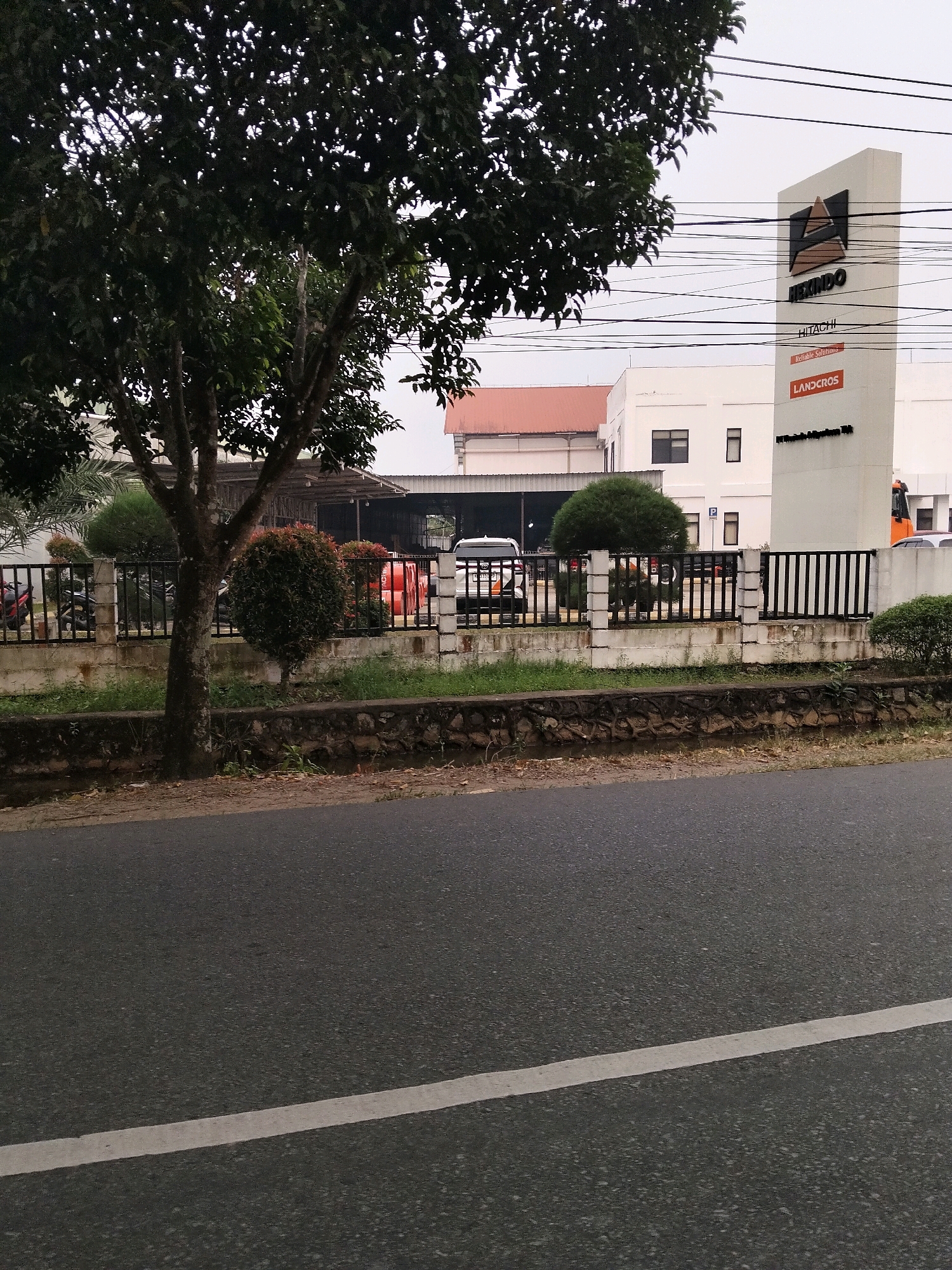 In Sungai Raya, Indonesia, a large tree with dark green foliage stands in the foreground on the left, partially obscuring the view of a building complex. In front of the complex is a black metal fence supported by concrete pillars. Beyond the fence, several parked vehicles, including motorcycles and a white van, are visible. A tall, white monument with dark lettering stands to the right of the building, featuring the logos "HINO" and "HITACHI." The monument has "Reliable Solutions" and "LANDGROS" printed in smaller text. A street with a white dividing line runs across the bottom of the image. The sky is overcast.