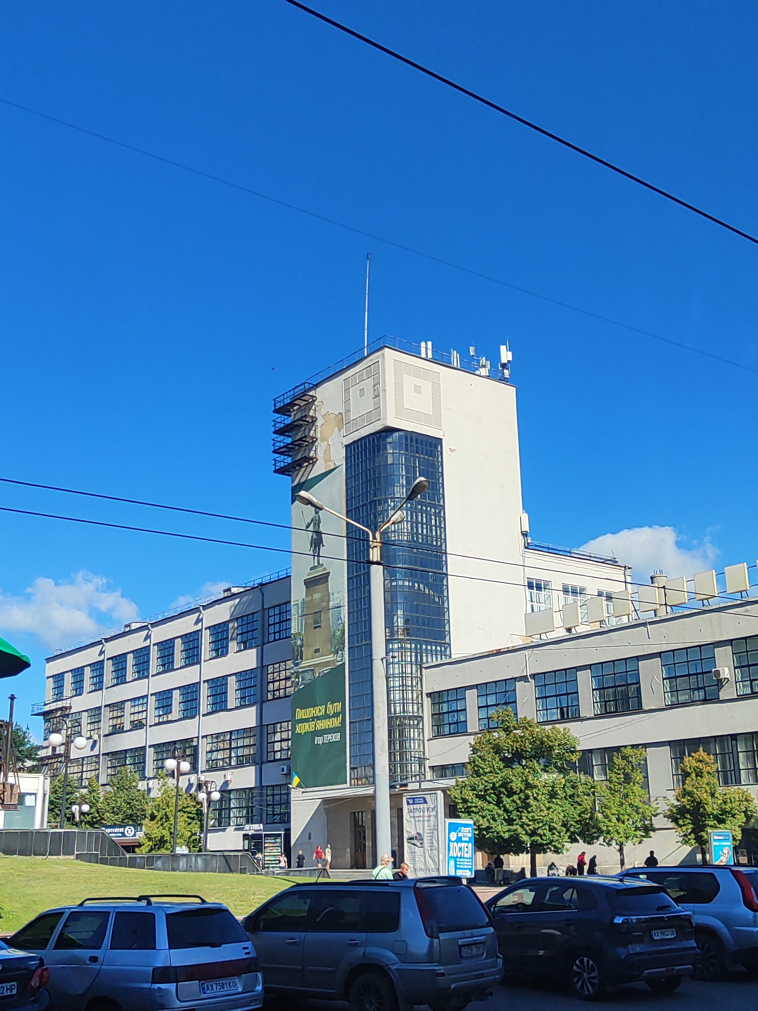 The image shows a multi-story, modern building with a prominent tower section, set against a bright blue sky with scattered white clouds. The building features large glass windows and a mural depicting a statue on a pedestal. Various antennas are visible on the rooftop of the tower. In the foreground, a grassy slope leads down to a street where several cars are parked, including silver sedans and SUVs. People are walking on the sidewalk near the building entrance. The scene appears to be in an urban environment during daylight hours.