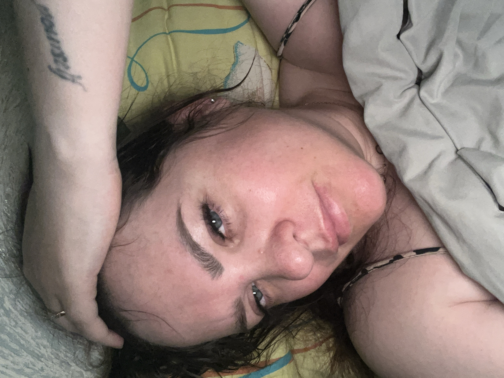 A woman lies on a yellow and teal patterned bed with a grey blanket partially covering her body. She is fair-skinned with brown hair. Her left arm is raised above her head, exposing a tattoo on her inner forearm. A ring is visible on her left ring finger. She has grey eyes and is looking at the camera. A small piece of jewelry is visible in her left ear. A necklace is around her neck. The location is not specified in the image, only mentioned in the prompt as Noviy, Russia.