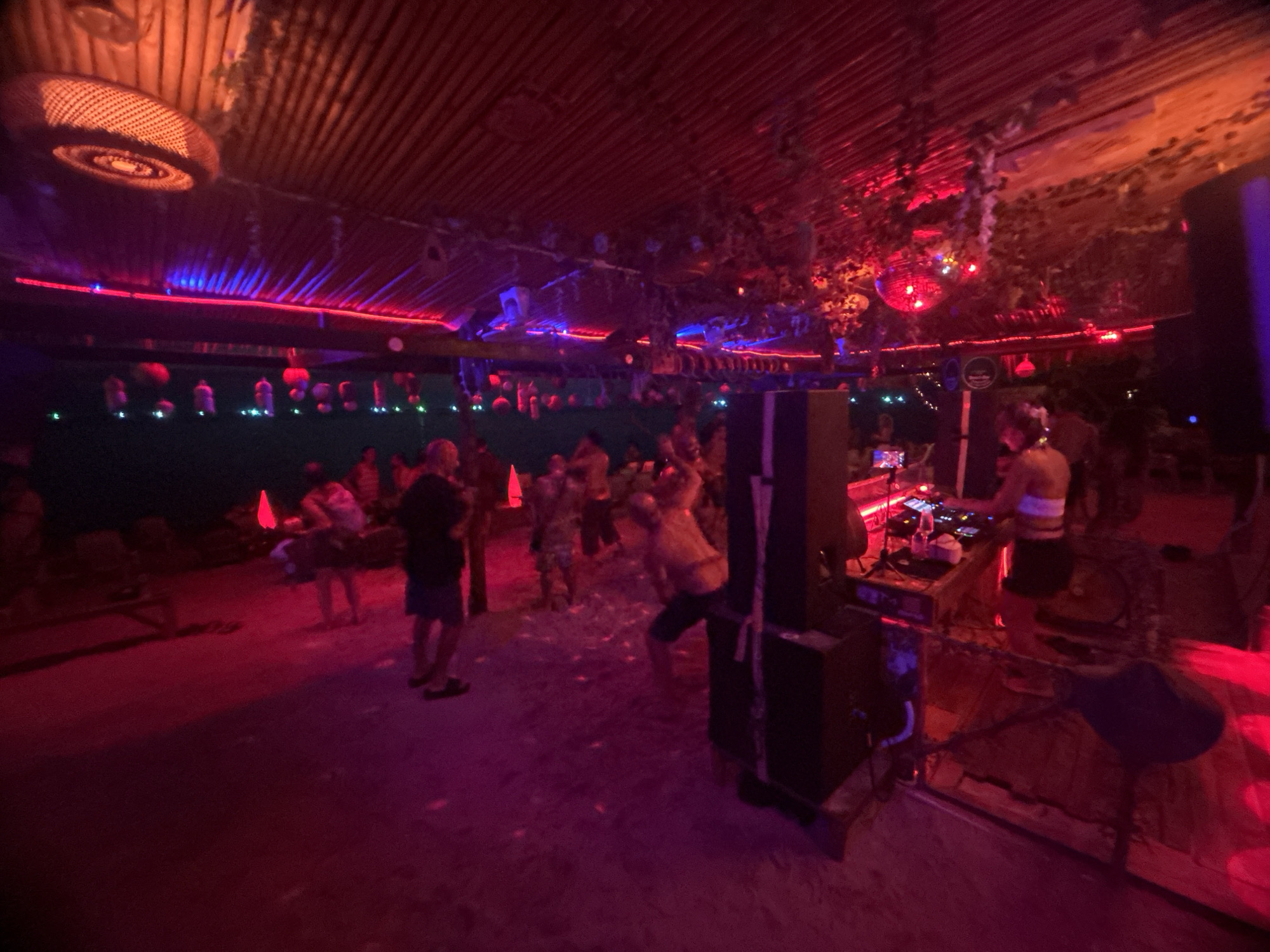 Here is a description of the image:
The image shows a nightlife scene at a venue in Baan Sri Thanu, Thailand. The building has a thatched roof with red and blue lighting fixtures. People are gathered, some appear to be dancing, in an area with sand on the ground. In the foreground is a DJ booth where a woman is operating equipment. Speakers are placed on either side of the booth. The overall lighting is dim and colored red and purple. The environment has vegetation hanging down in places.